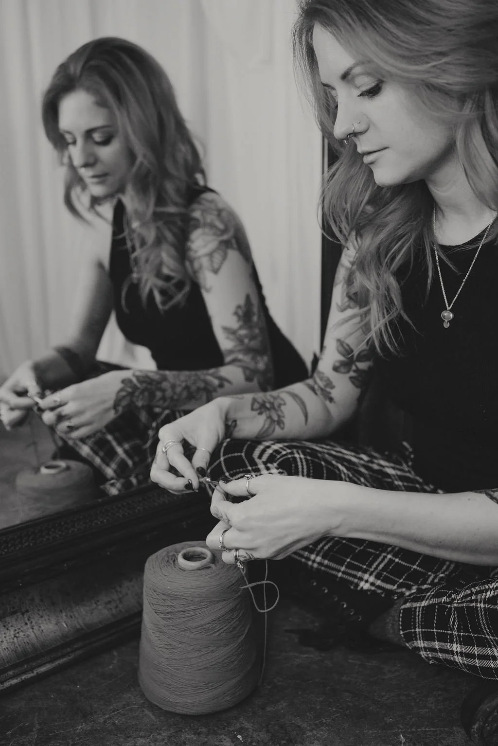 A woman with tattoos and jewelry, sitting cross-legged, knitting or working with a spool of yarn in front of a mirror.