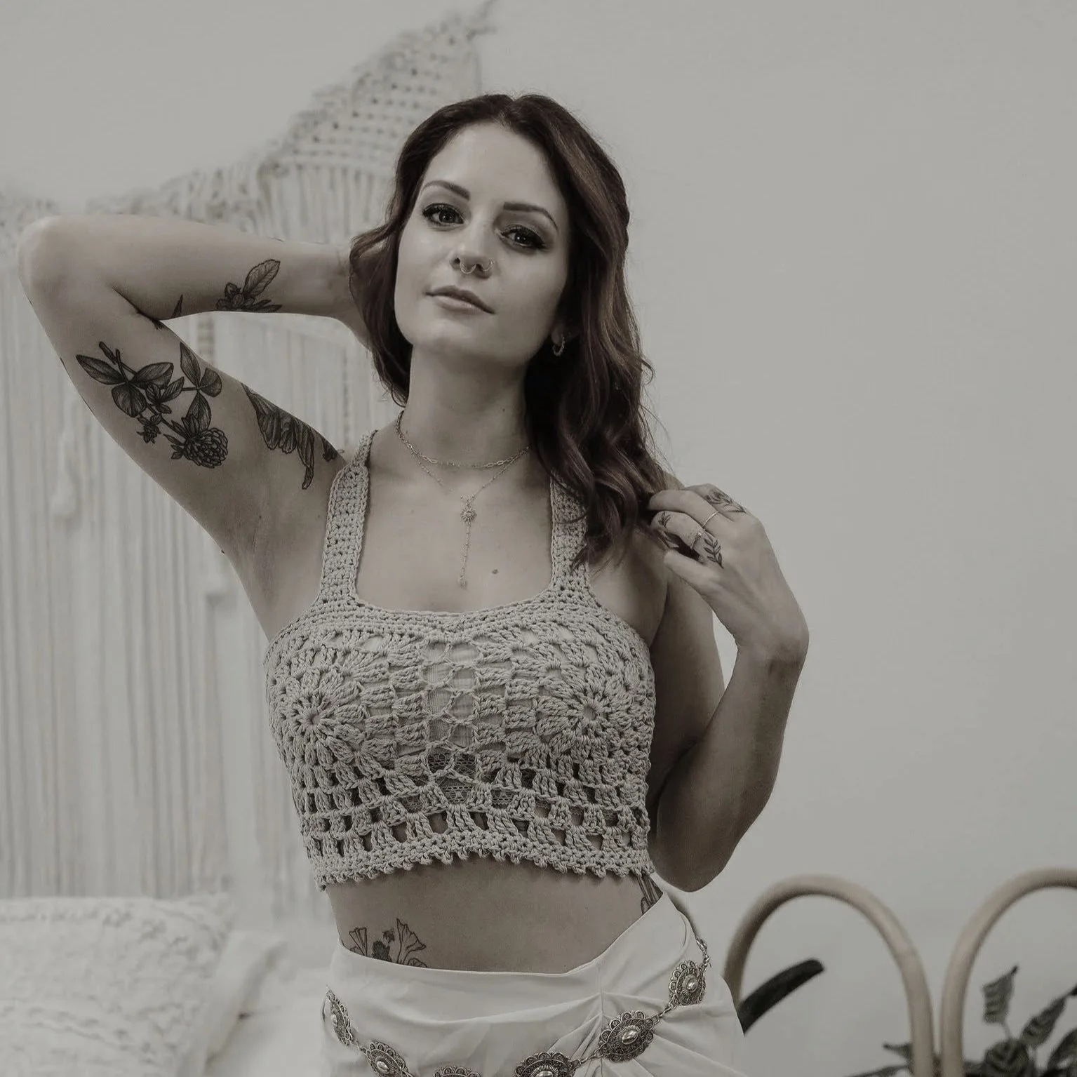 A young woman with tattoos on her arm and side, wearing a crocheted crop top and light-colored skirt, posing in front of a mirror.