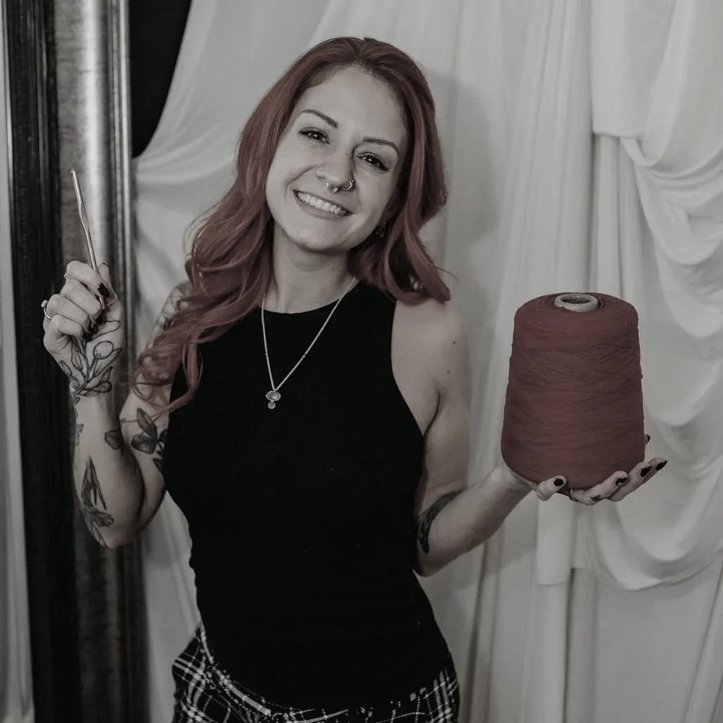 A woman with red hair and tattoos holding a spool of red thread and a needle, smiling at the camera.