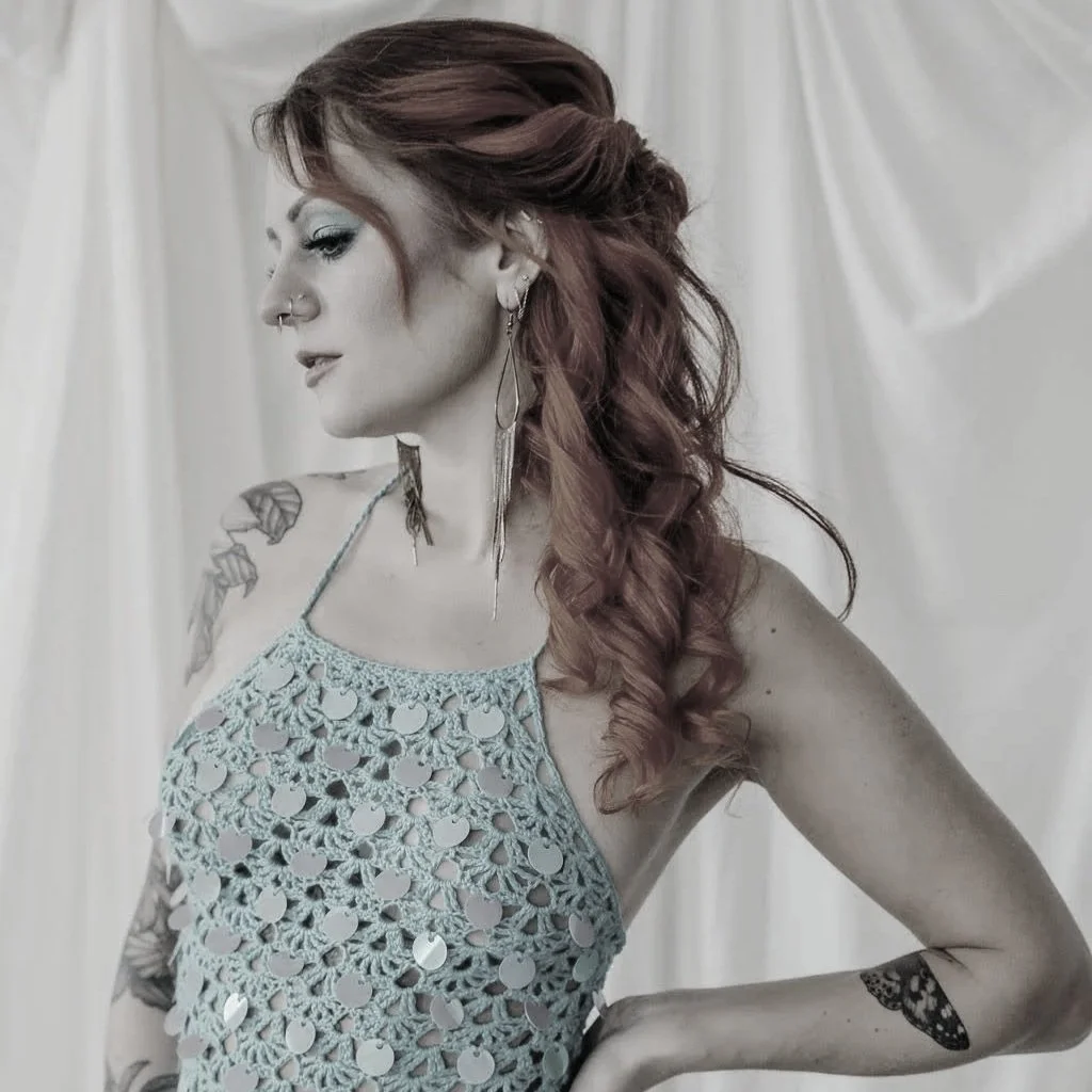 A woman with red hair styled in loose curls, wearing a crocheted halter top, earrings, and tattoos on her arms, standing against a light-colored curtain backdrop.
