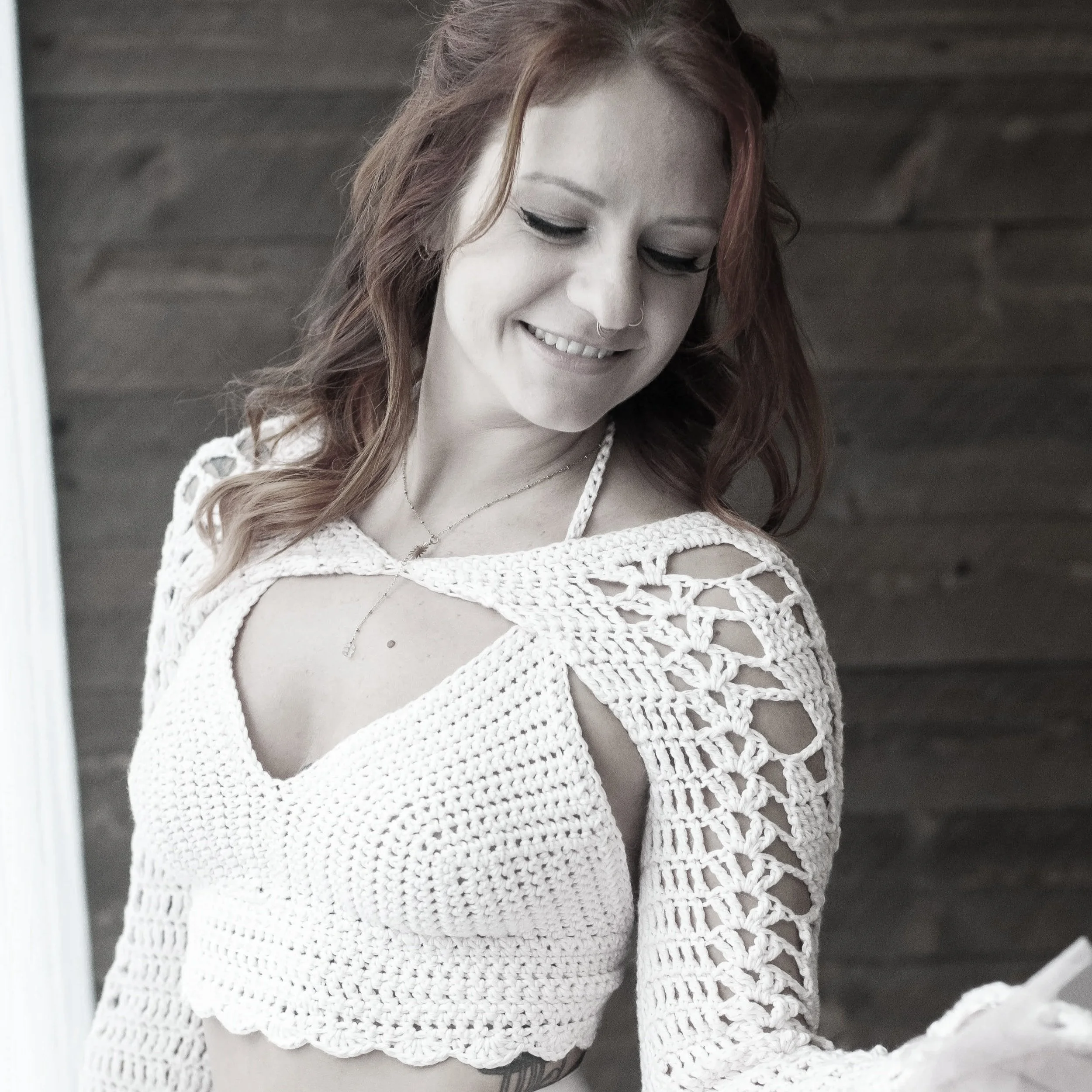 A smiling woman with red hair in a crochet crop top with open shoulder details, looking down. She is standing in front of a wooden wall.