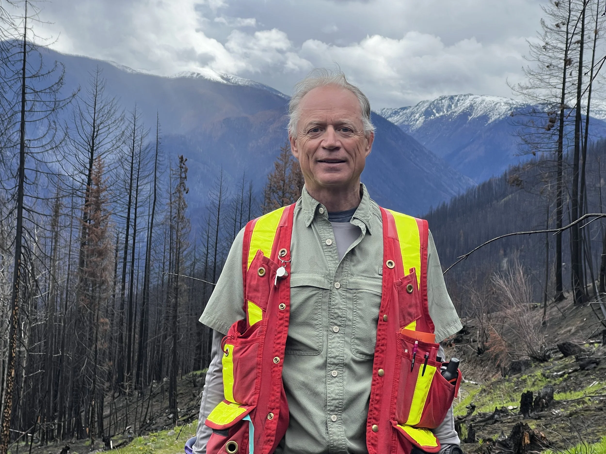 Mike Madill standing outdoors in a burnt forest with mountain ranges in the background, wearing a gray shirt and a bright yellow and red safety vest.