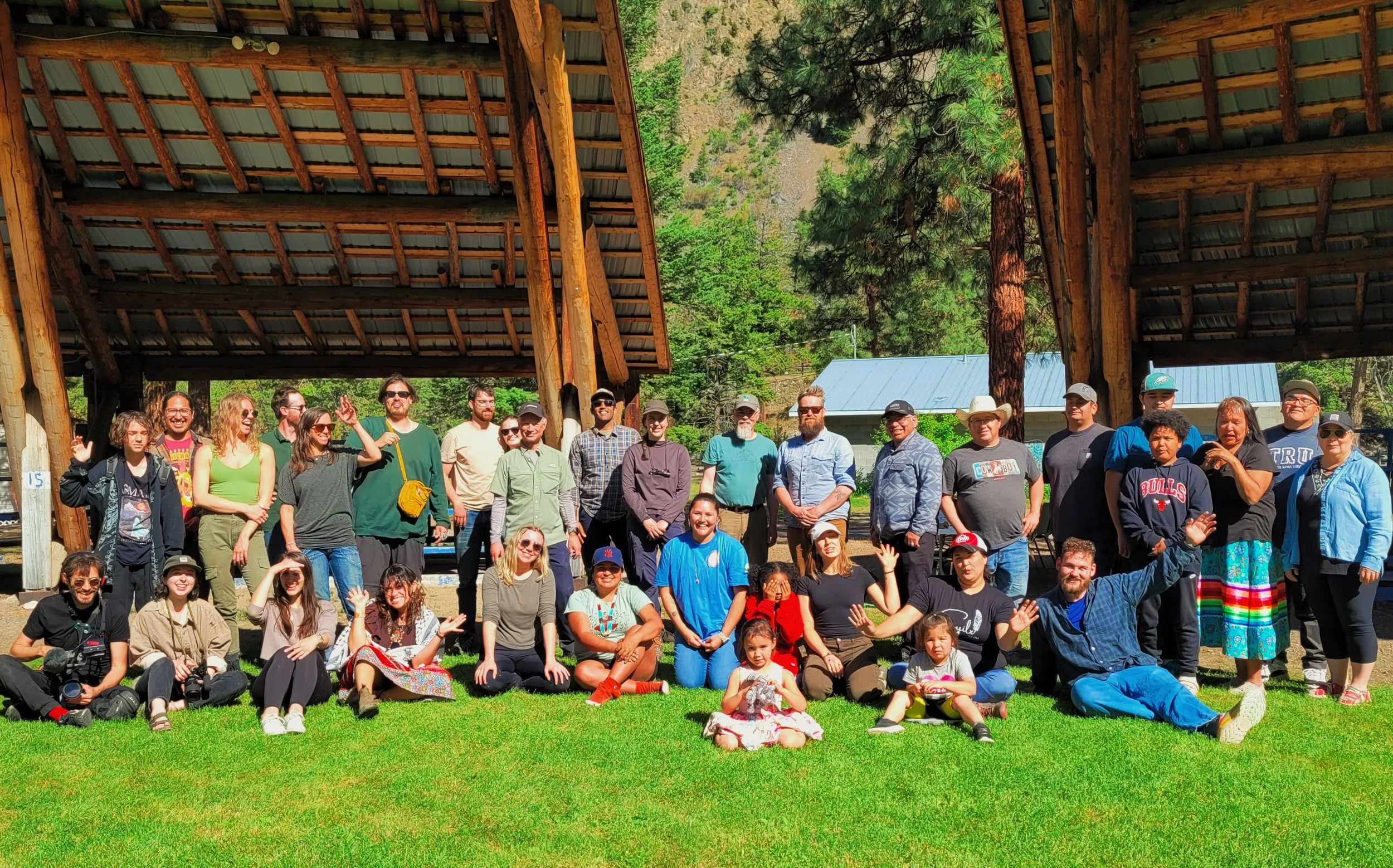 Group photo of diverse people including First Nations communities, gathering outdoors, standing and sitting on grass under wooden shelter in a forested area on a sunny day.