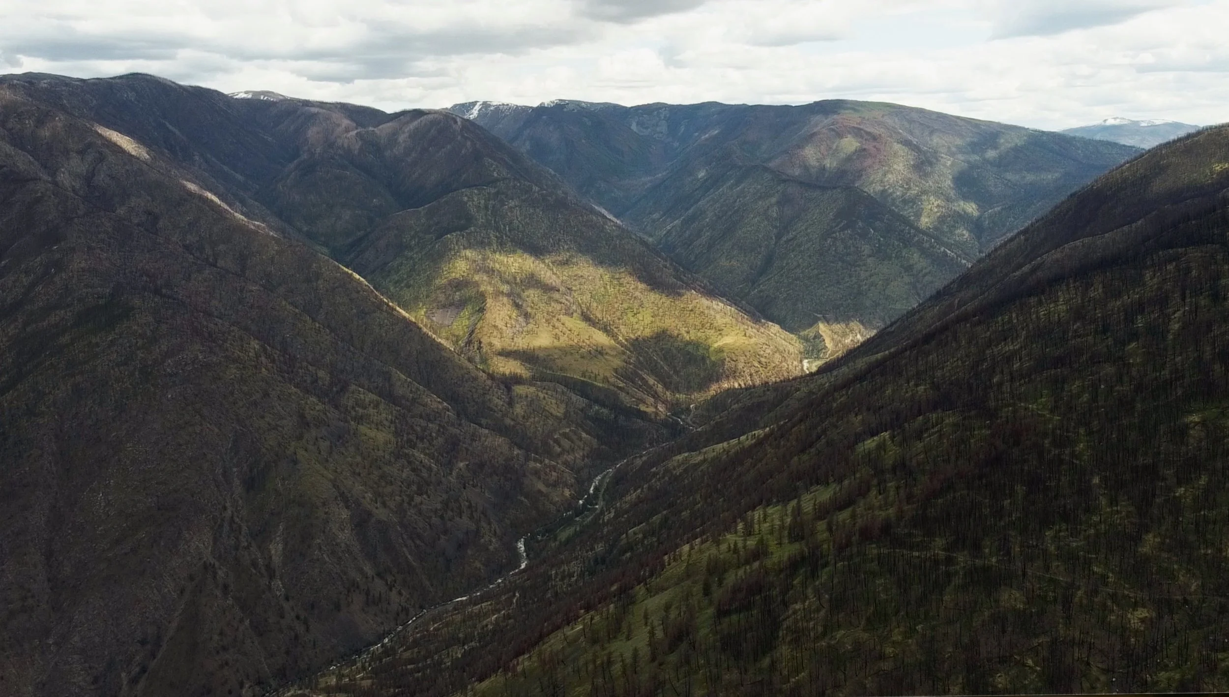 Mountain landscape with green and brown slopes, some snow patches on the peaks, and a cloudy sky.