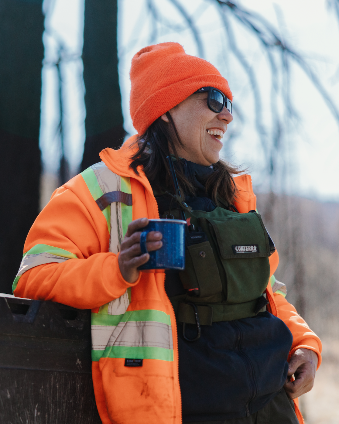 Jeni Christie wearing a bright orange beanie and jacket, holding a blue mug, smiling outdoors in a wintery forest setting.