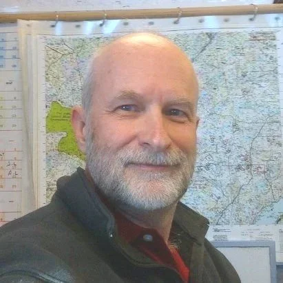 Mark Seilis with a beard and a bald head, smiling, standing in front of a large map on the wall.