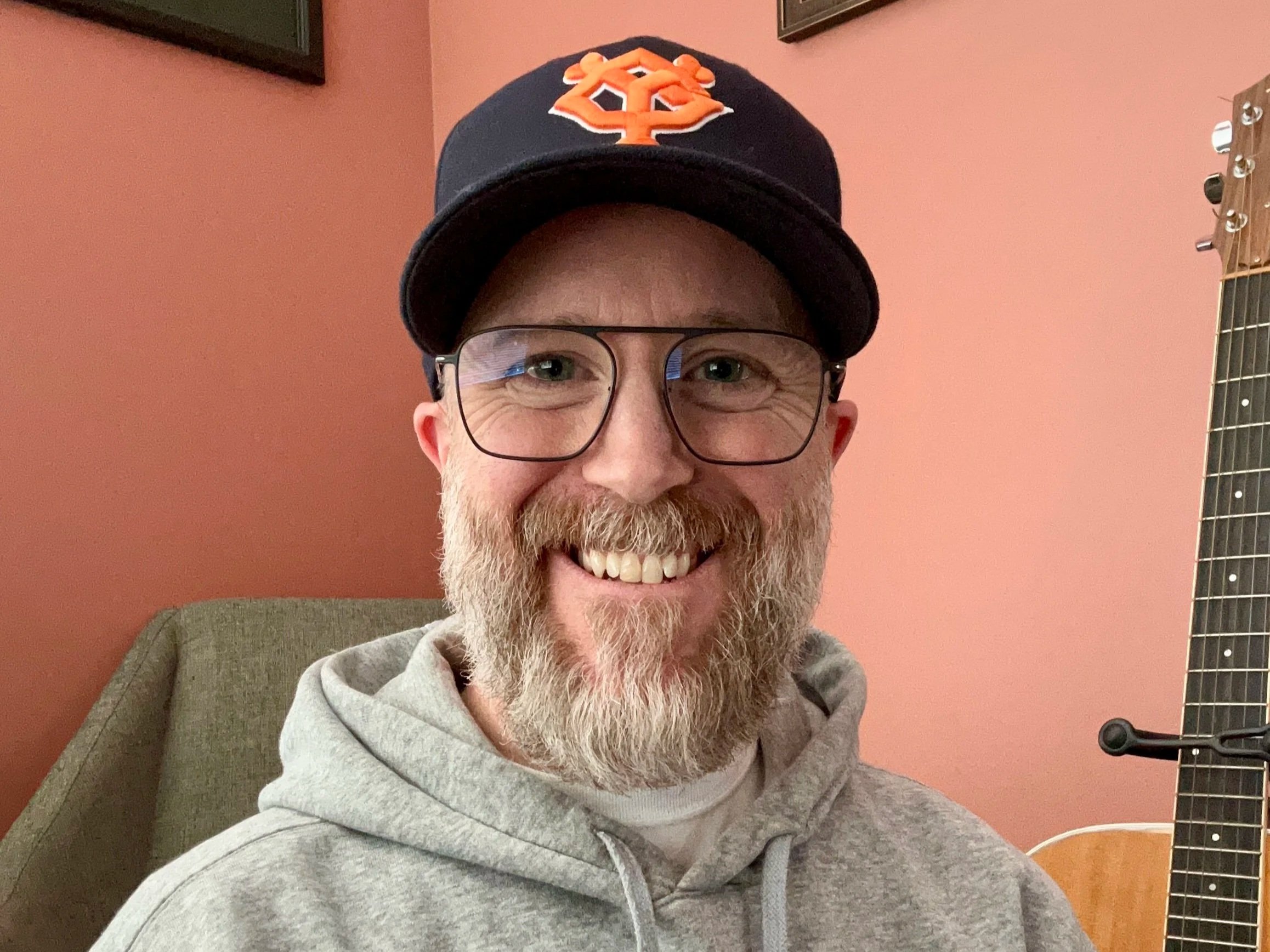 Wesley Brookes smiling, with a beard and glasses wearing a black baseball cap with an orange logo, a gray hoodie, and sitting next to an acoustic guitar in a room with pink walls.