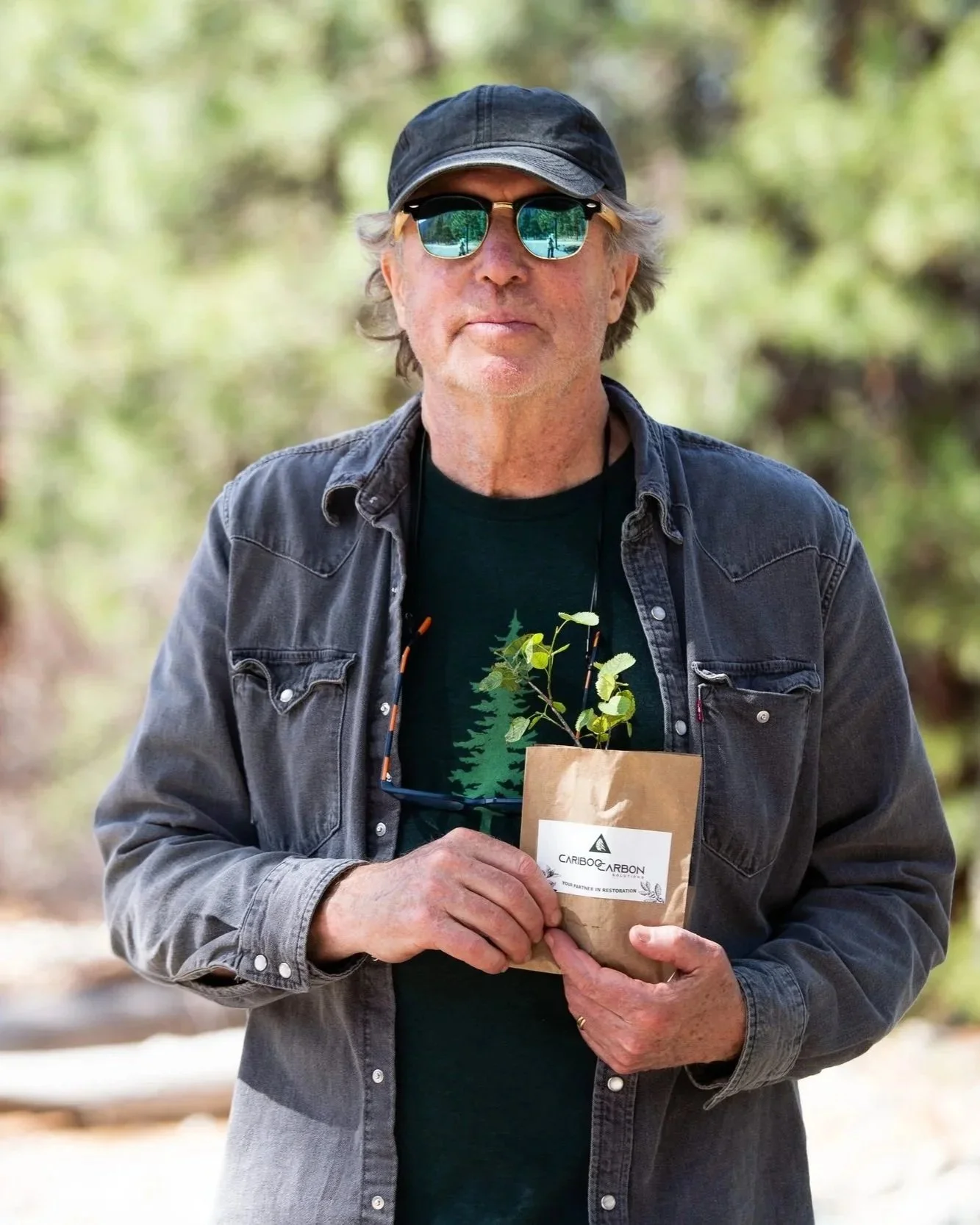 Tony Harrison wearing sunglasses, a black cap, and a denim jacket stands outdoors holding a plant in a paper bag with trees in the background.