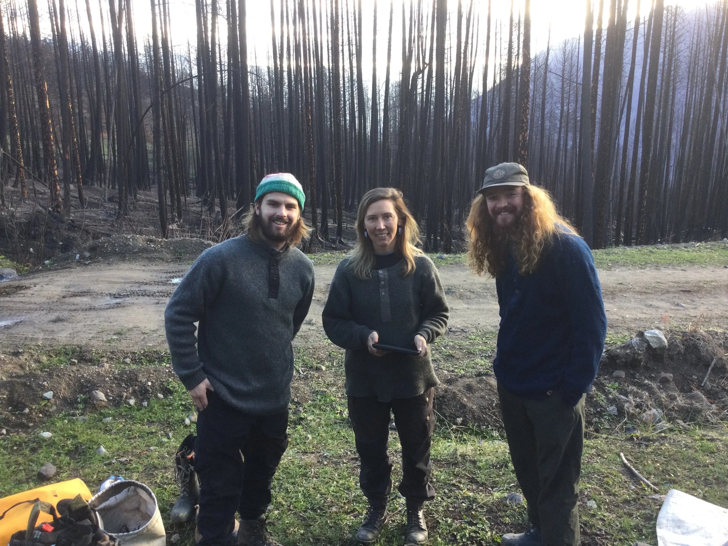Three people standing outdoors in front of a burned forest, smiling at the camera. The person on the left has long hair, a beard, a gray sweater, and a green beanie. The person in the middle has shoulder-length hair, wearing a dark sweater and holding a tablet. The person on the right has long curly hair, a beard, a gray cap, and a dark jacket.
