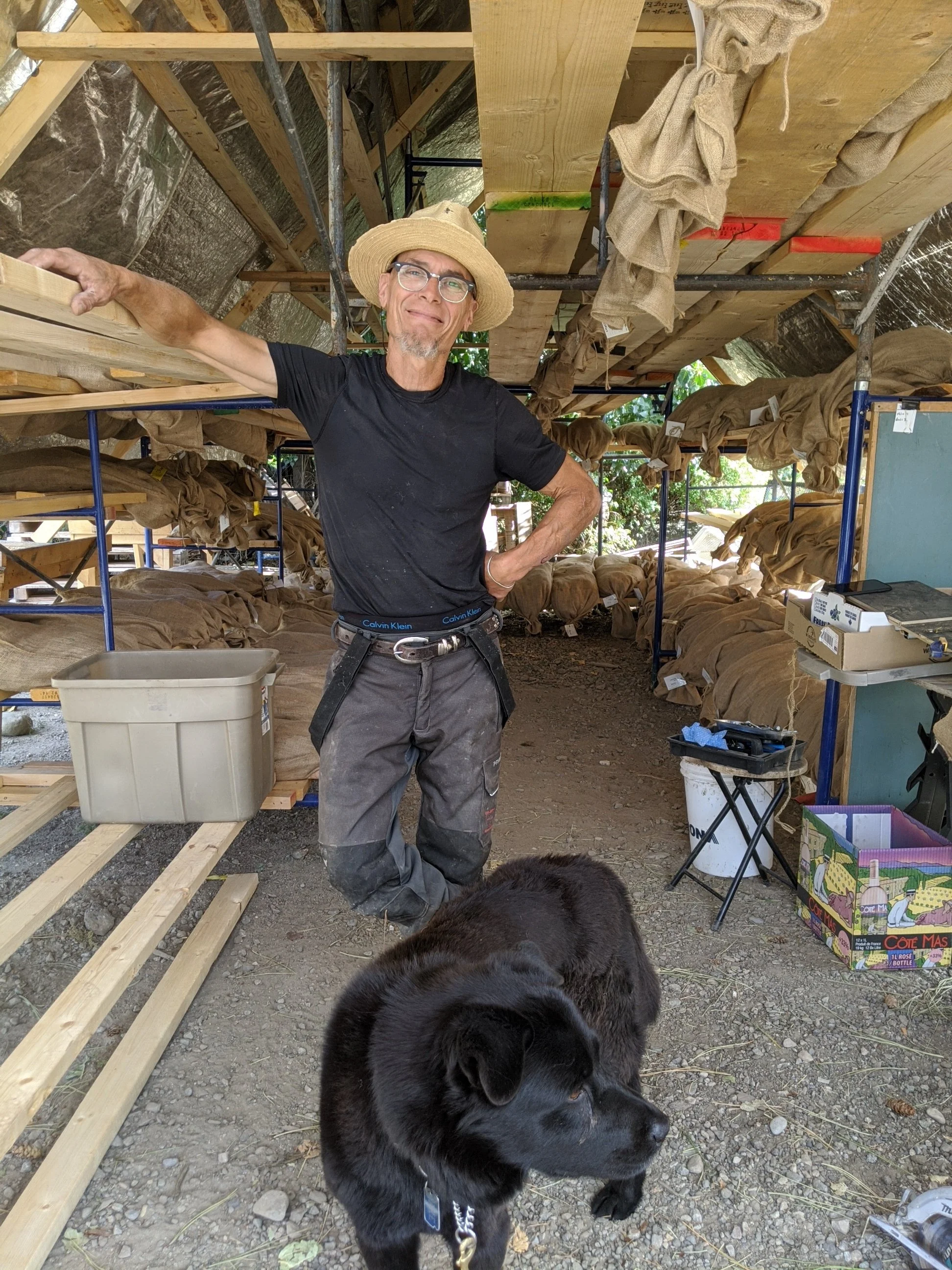 A man wearing a straw hat, black t-shirt stands inside with one hand on his hip A black dog is in the foreground. The shelter has wooden beams and scaffolding, with dried tree seeds, cones and various tools and supplies visible.