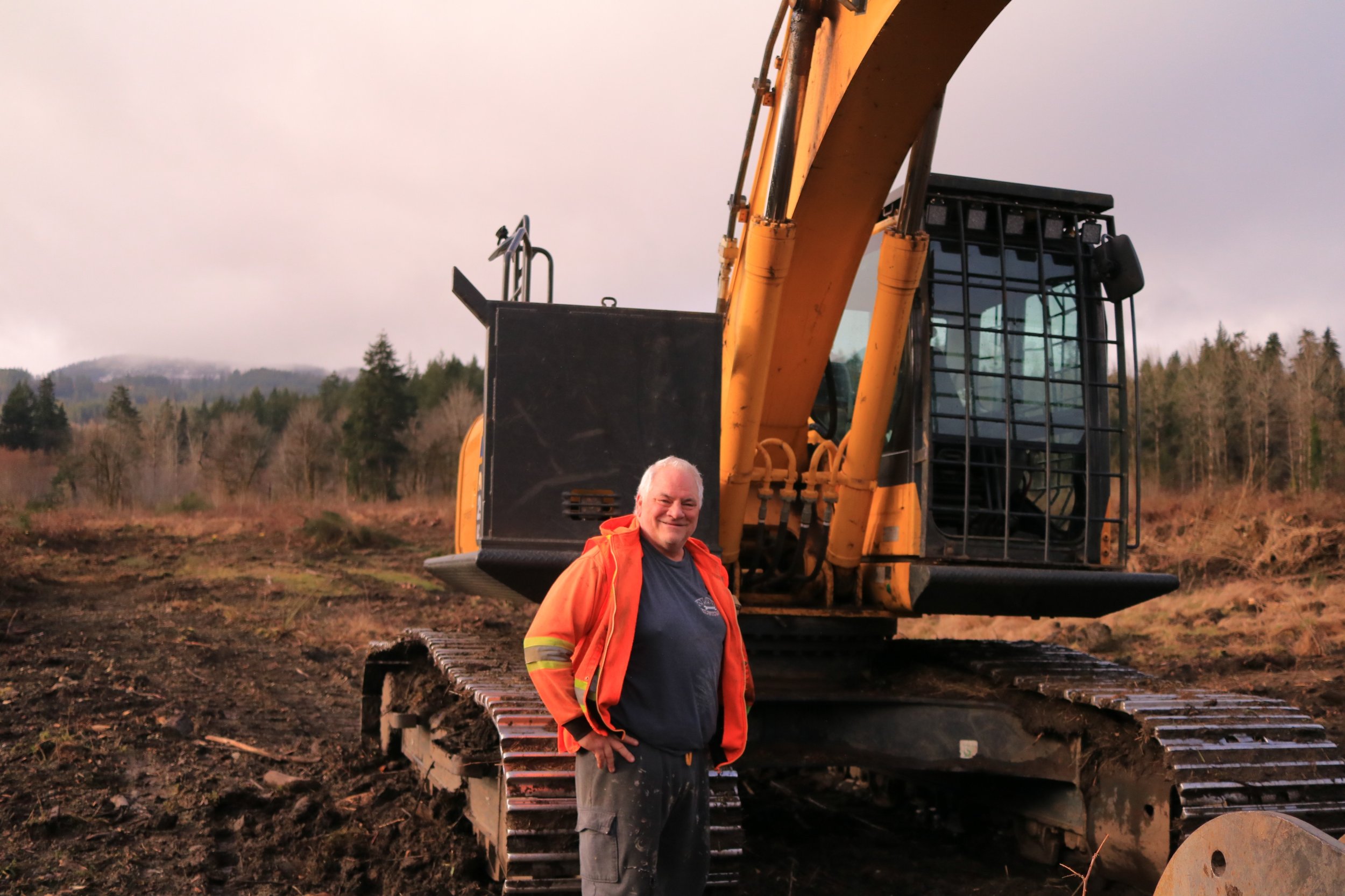 A smiling man standing in front of a large yellow excavator on a construction site, with dirt and a forested background.