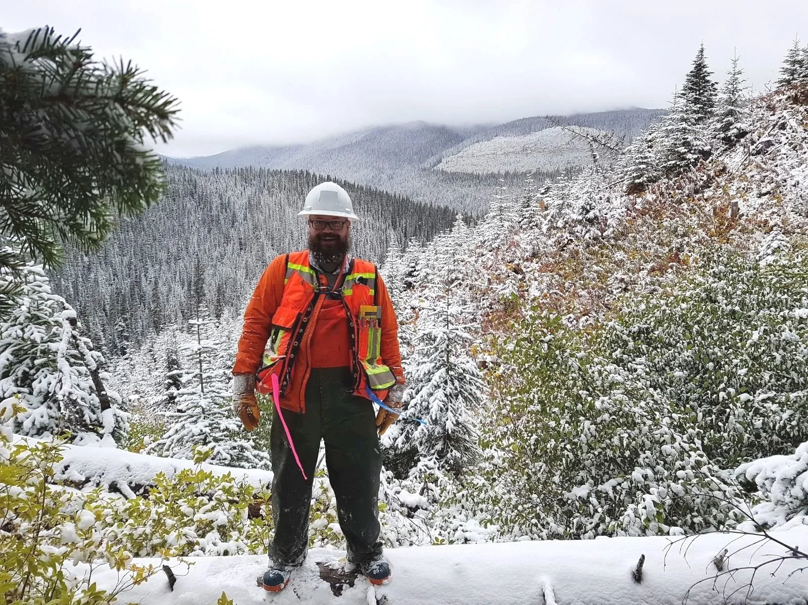 Justin Morrison wearing a white safety helmet, orange jacket, and safety vest stands in a snowy forested mountain landscape.