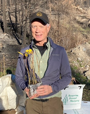 Mike Madill outdoors holding a clear container with yellow flowers and brown stems, wearing a dark cap and jacket, with a background of leafless trees and rocky terrain.