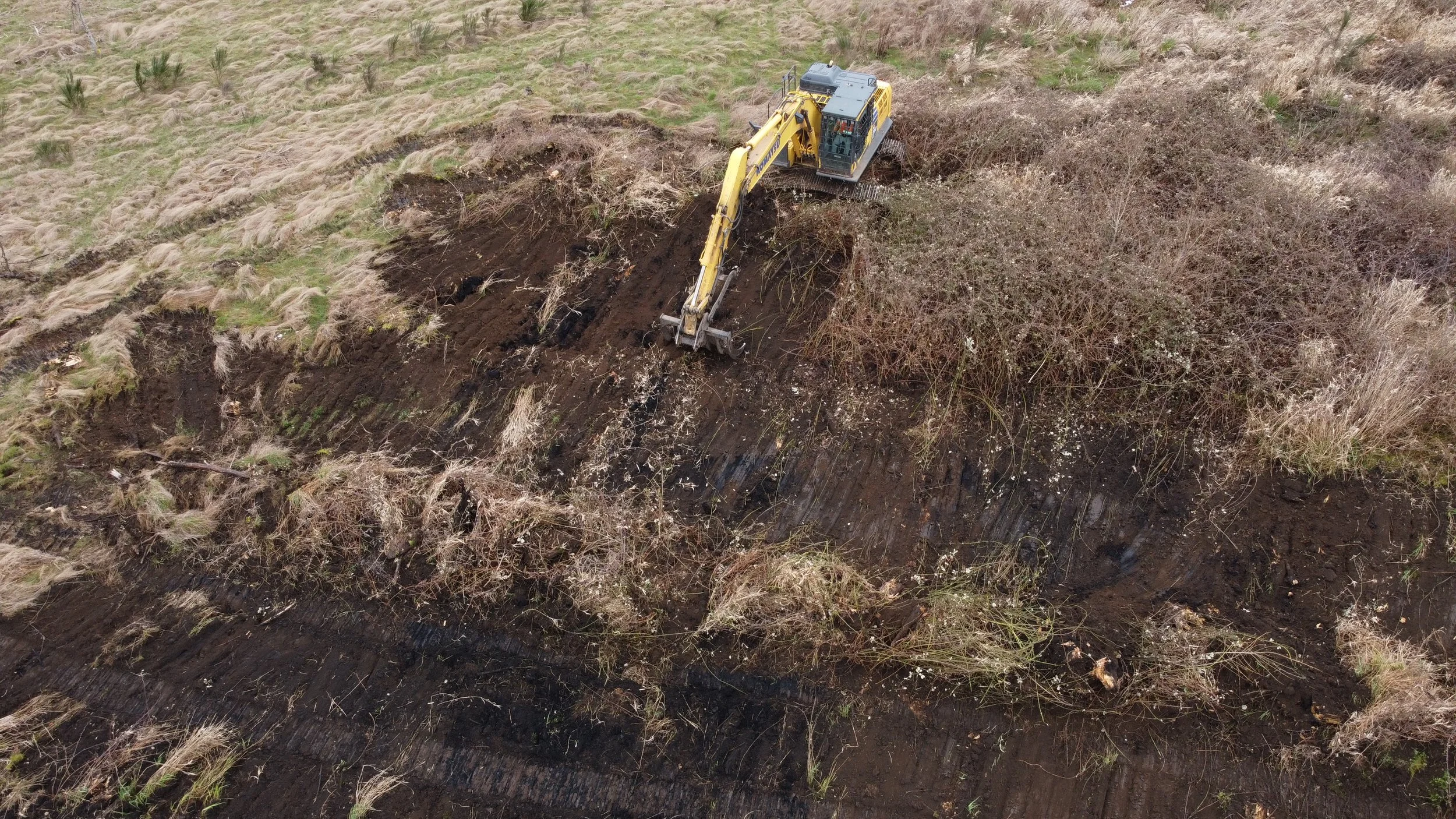 A yellow excavator working on the side of a hill, excavating soil and vegetation during a land clearing process.