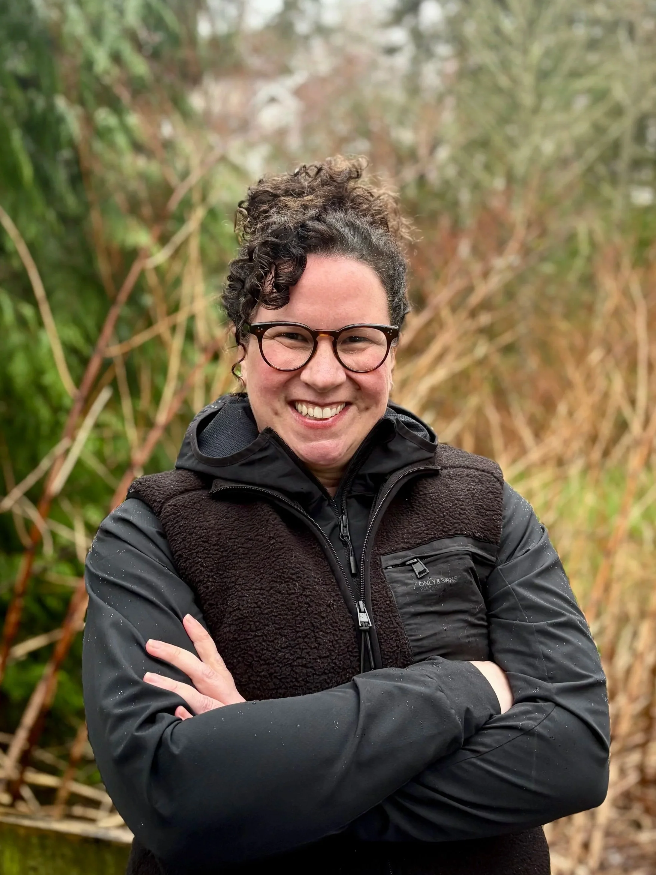 Mia Finn, in glasses, and a black outdoor jacket, smiling with arms crossed, standing outdoors in front of a background of trees and shrubs.
