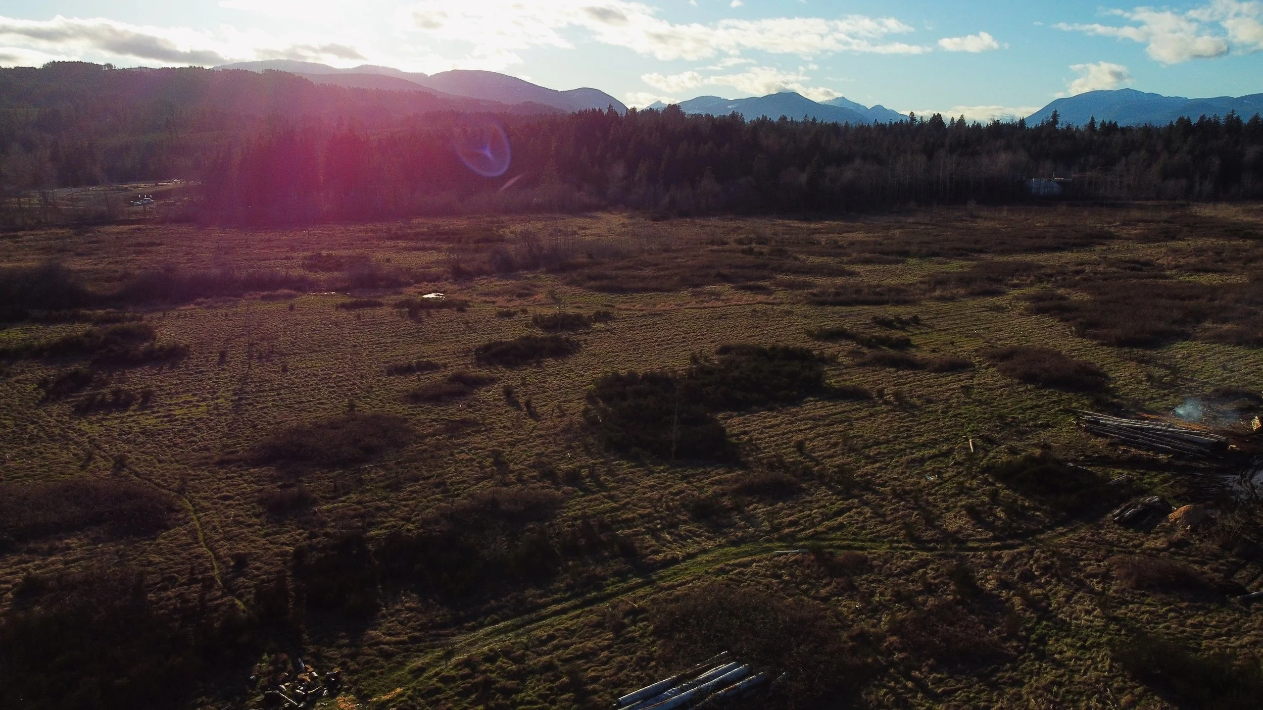 Aerial view of a rural landscape with fields, trees, and mountains in the distance under a partly cloudy sky with the sun shining.