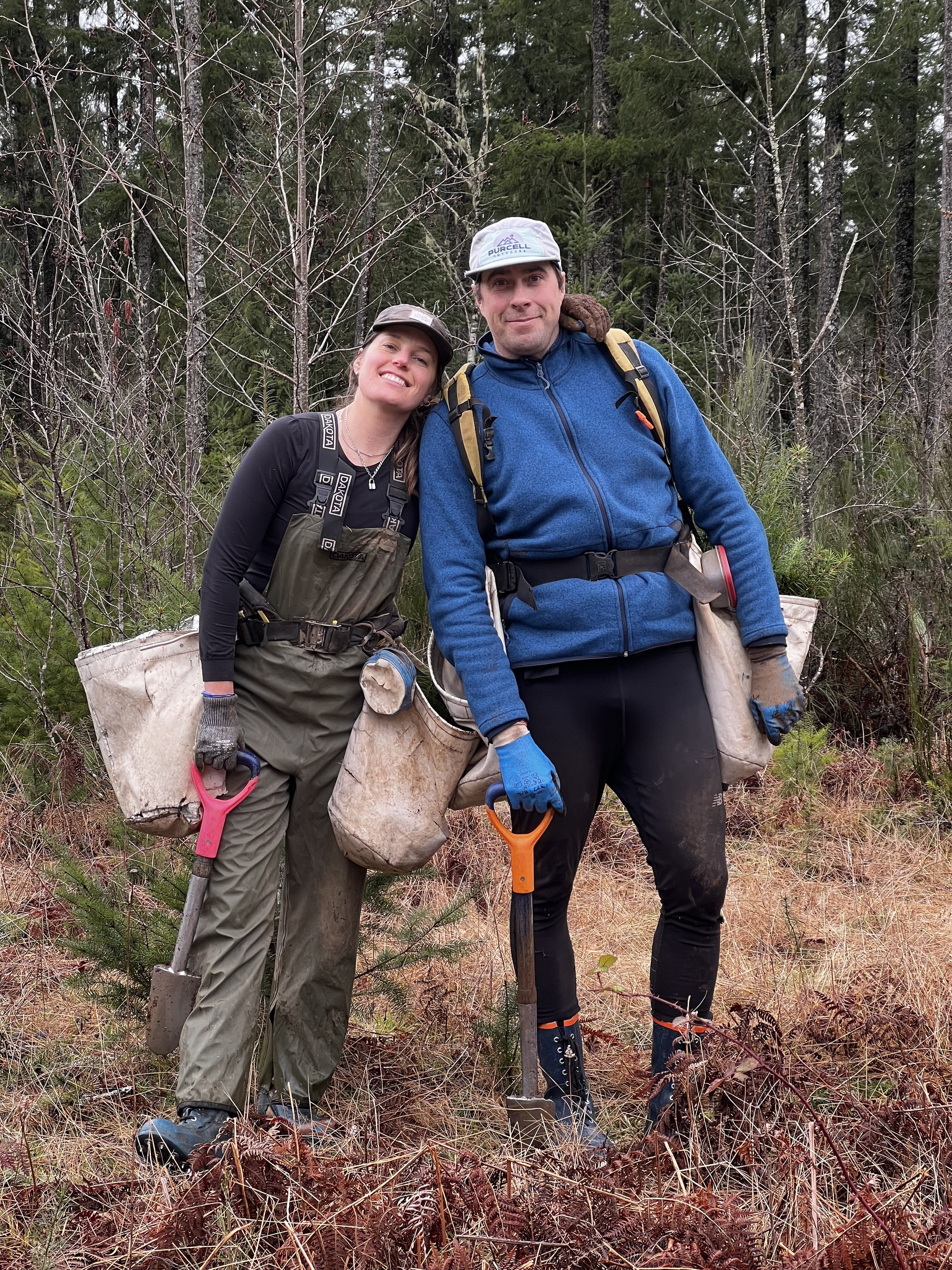 Two people standing in a forested area with leafless trees and grassy ground, dressed for outdoor work or exploration, holding small shovels and wearing backpacks and gloves.