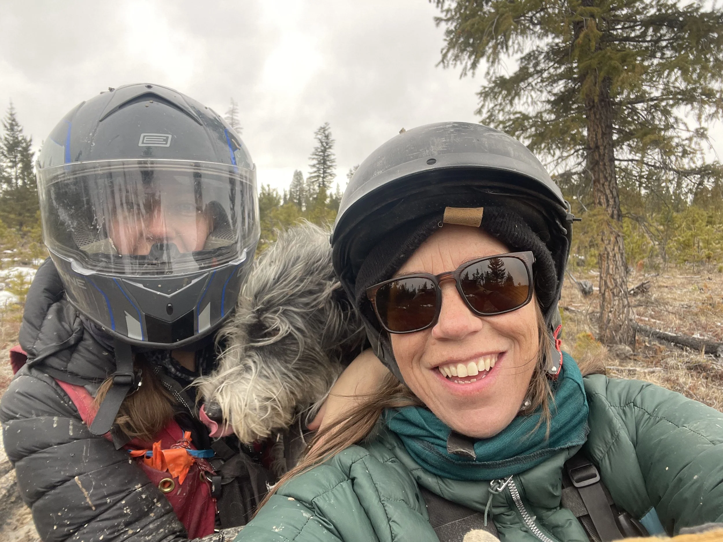 Two women wearing motorcycle helmets and outdoor jackets, one smiling and the other partially visible, with a dog nestled between them, in a wooded outdoor setting.