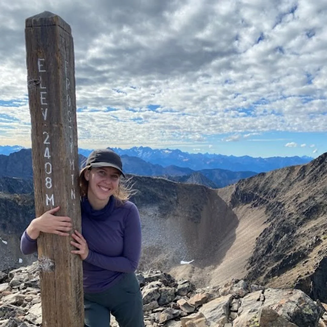 A woman stands next to a wooden signpost at the summit of a mountain, with the crater of a volcano behind her. The sign reads 'Elevation 2408 M'. She is smiling, wearing a purple long-sleeve shirt and a gray hat. The background features rugged mountain peaks and a partly cloudy sky.