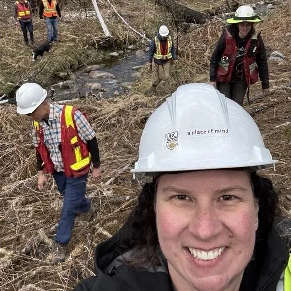 Mia Finn in a construction helmet smiling, surrounded by several people wearing safety gear and helmets operating in a natural outdoor setting with dry grasses and a small stream.