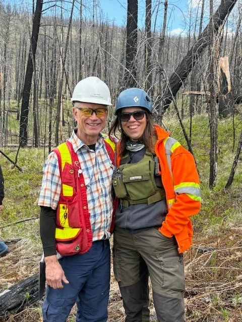 Mike and Jeni with hard hats on with wildfire trees and forest burned in the background