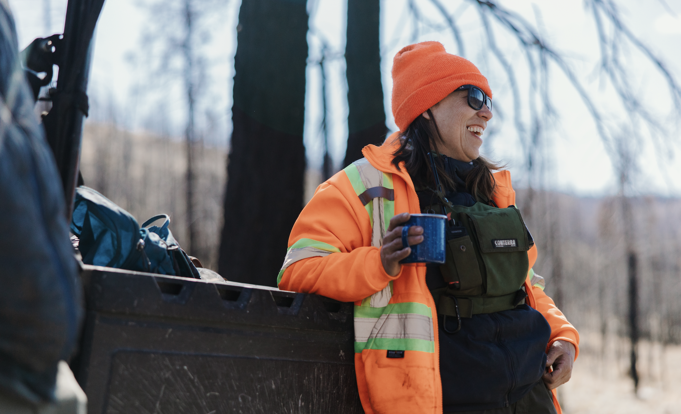 A woman in an orange safety jacket and bright orange beanie hat, wearing sunglasses, is smiling and holding a blue mug while leaning against a black truck in a winter forest.