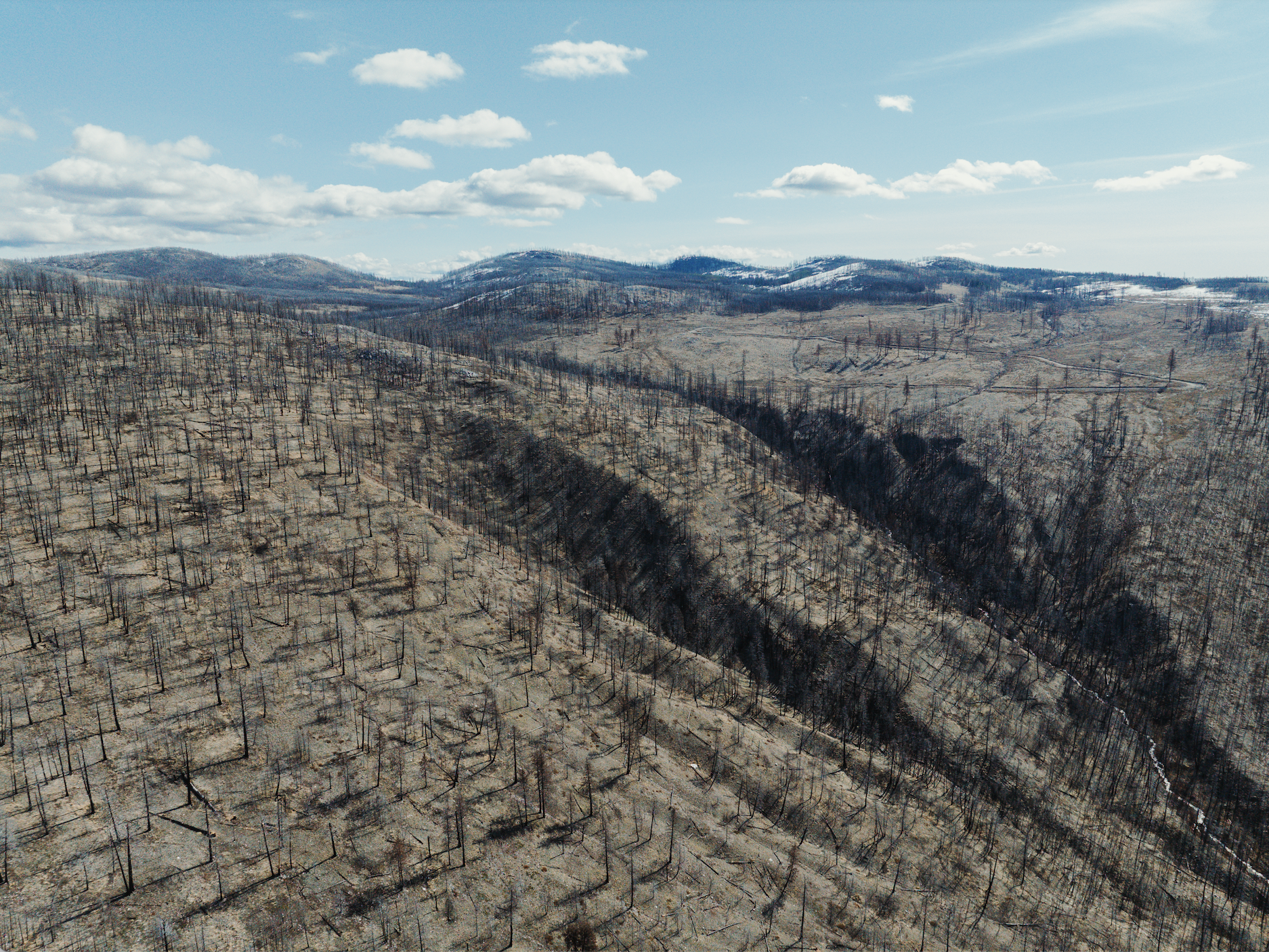 Aerial view of a mountain landscape with burnt trees and patches of snow, under a blue sky with scattered clouds.
