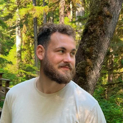 A young Mack with a beard and short, curly hair standing outdoors near a large tree in a forest, wearing a light-colored t-shirt and looking to the side.