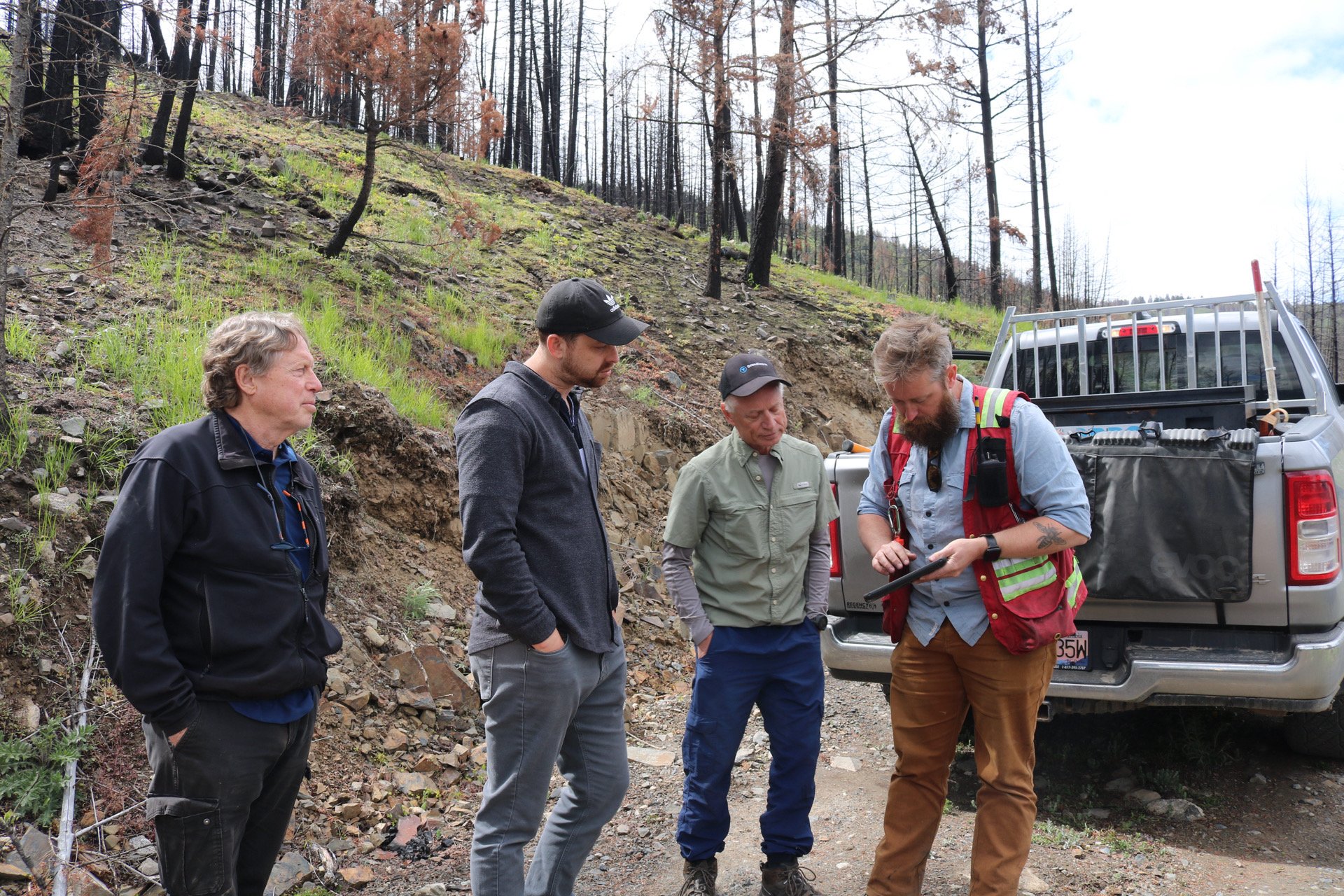 Four men standing outdoors in front of a burnt hillside, looking at a tablet held by one of the men, with a pickup truck behind them.
