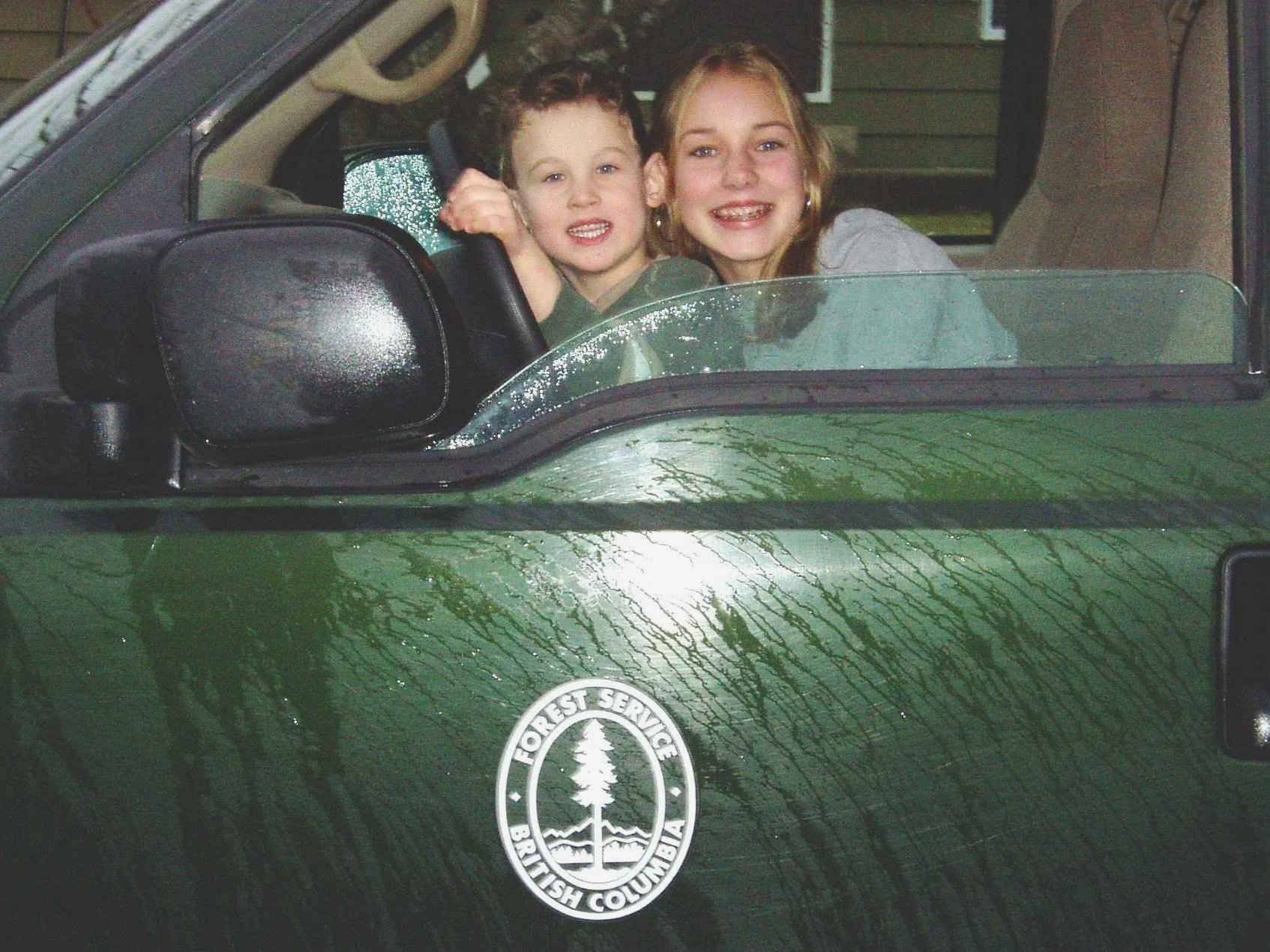 Young Mack and his sister as a boy and a girl, sitting inside a green vehicle with a Forest Service logo, smiling and looking out from the front, with the girl holding the steering wheel.