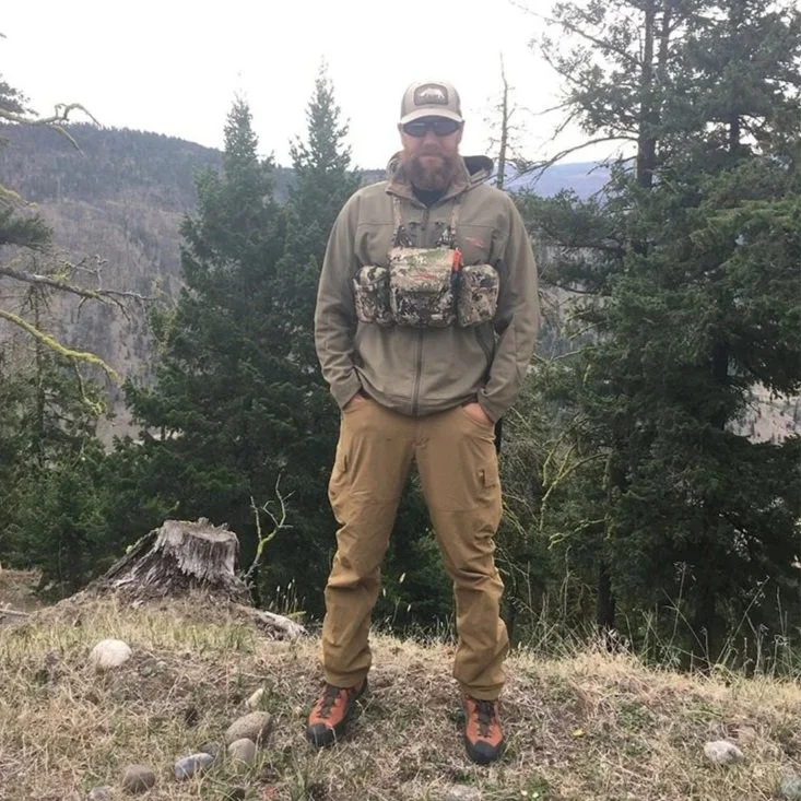 Andy Belicka in outdoor hiking gear standing on a grassy area with trees and mountains in the background.