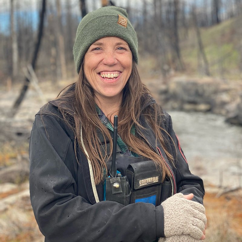 Jeni Christie, Senior Surveyor, smiling outdoors near a river, wearing a green beanie, a black jacket, beige gloves, and radio equipment.