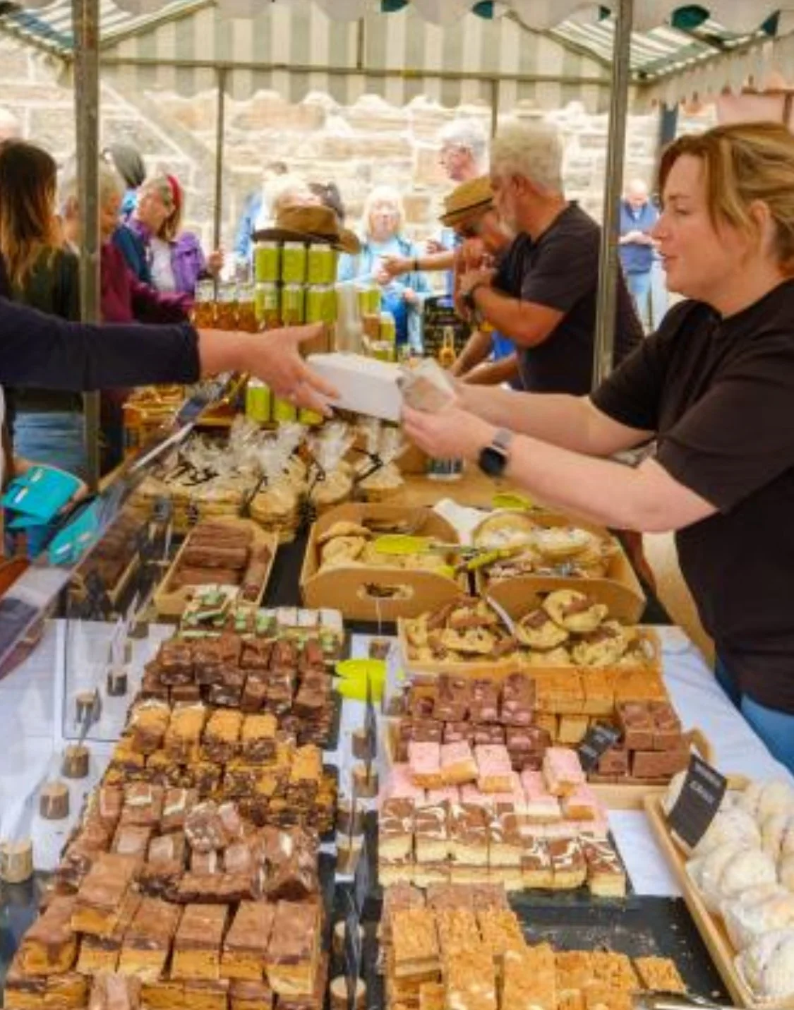 People shopping at an outdoor market stall with various baked goods and treats displayed on the table, and other shoppers seen in the background.
