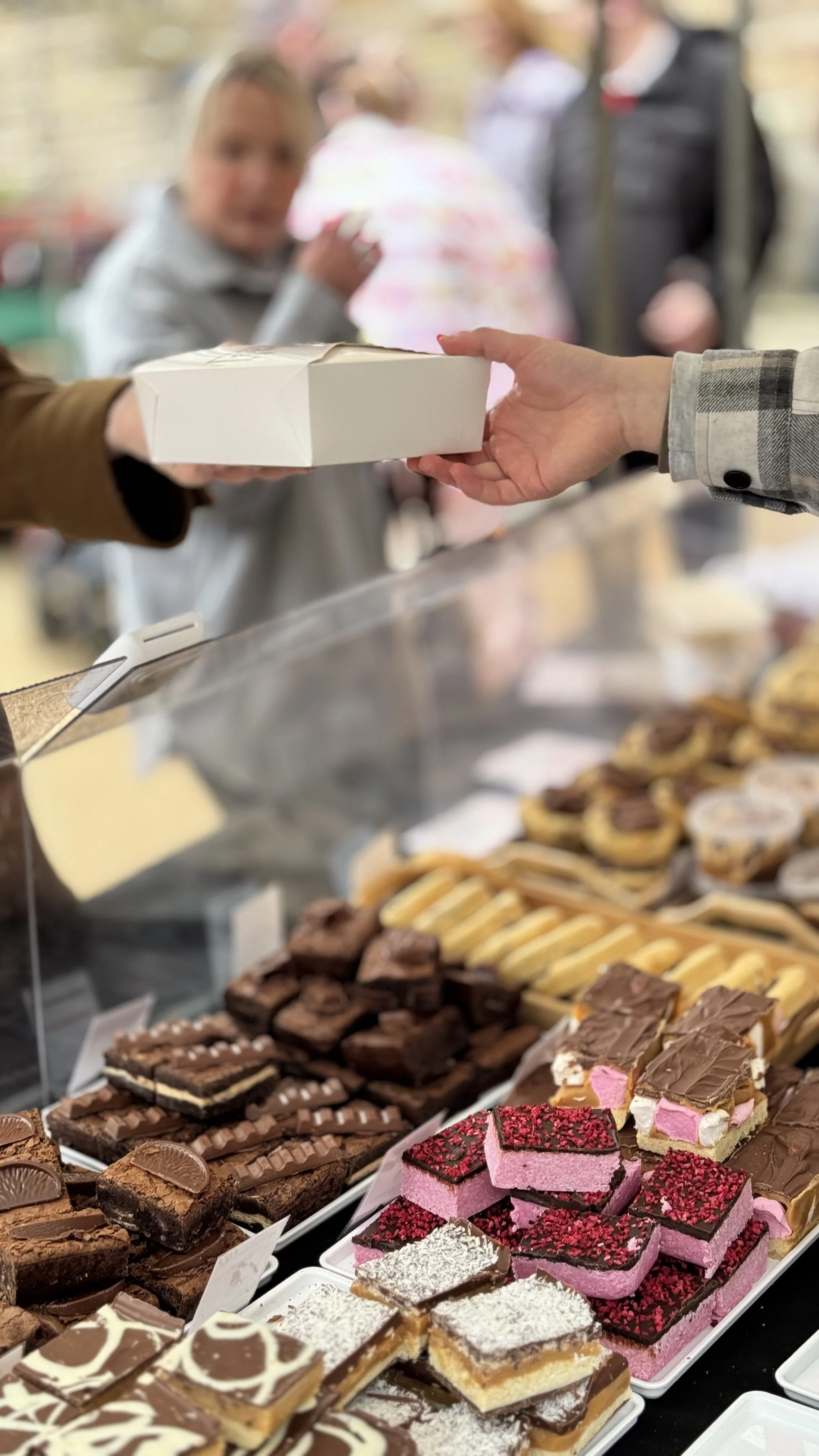A person is receiving a dessert box from another person at a bakery or cafe, with a display case of assorted baked goods including brownies, layered bars, and decorated cakes in the foreground.