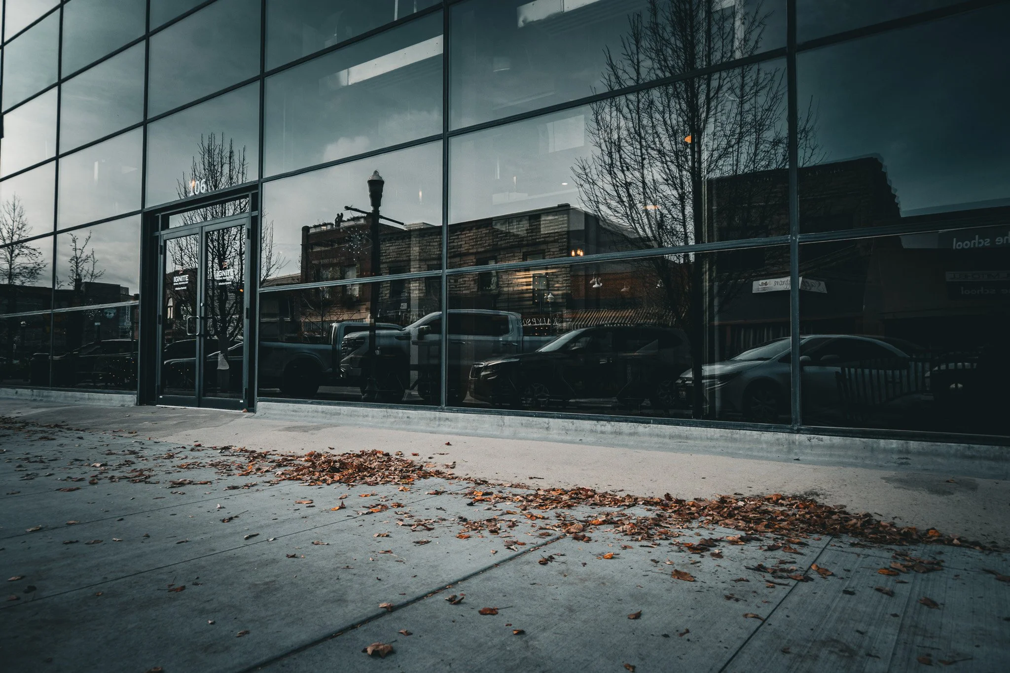 Reflection of parked cars, leafless trees, and buildings in a large glass window of a modern building, with fallen leaves on the sidewalk in the foreground.