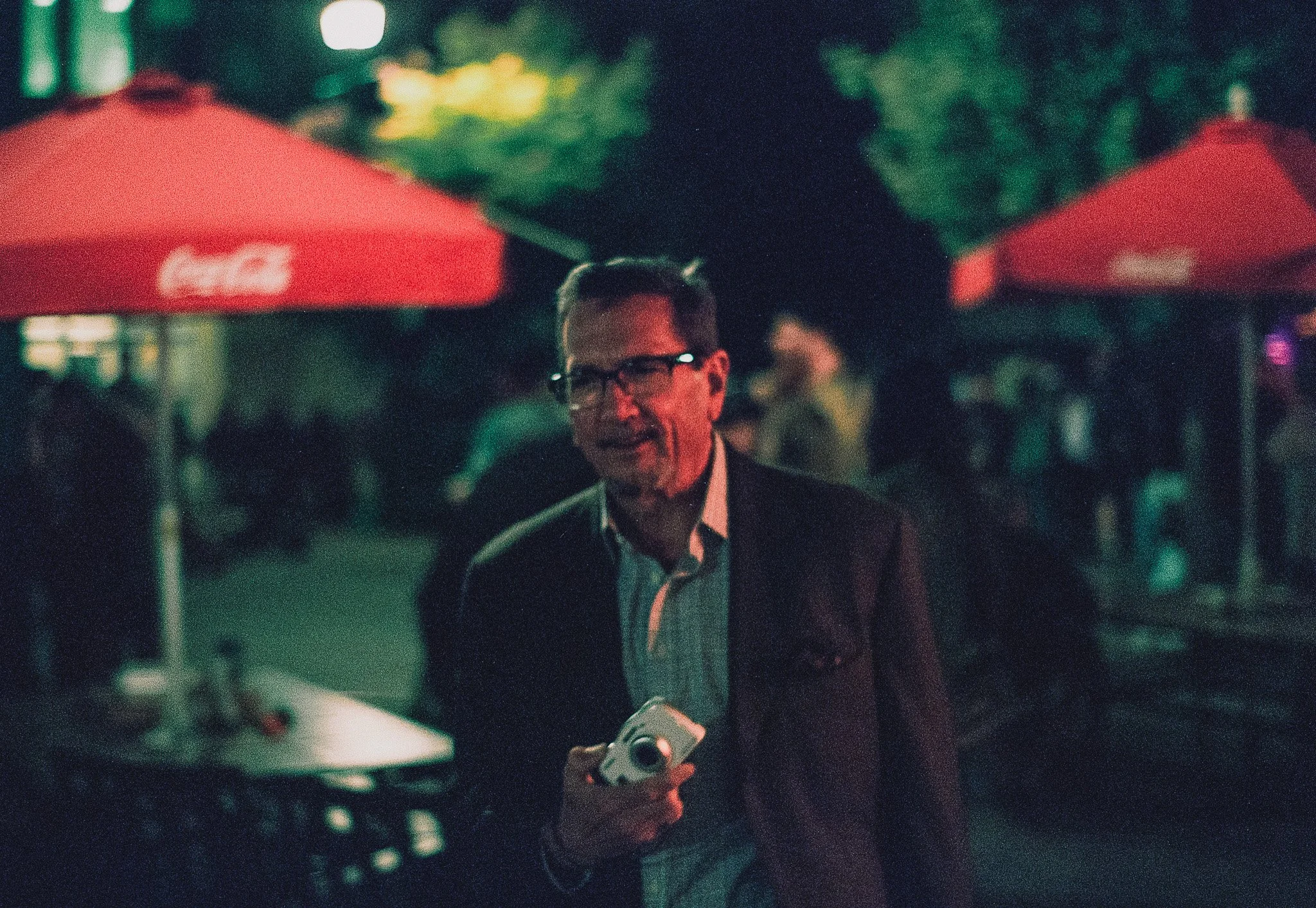 A man with glasses and a suit holding a camera at an outdoor event during nighttime, with red umbrellas and people in the background.