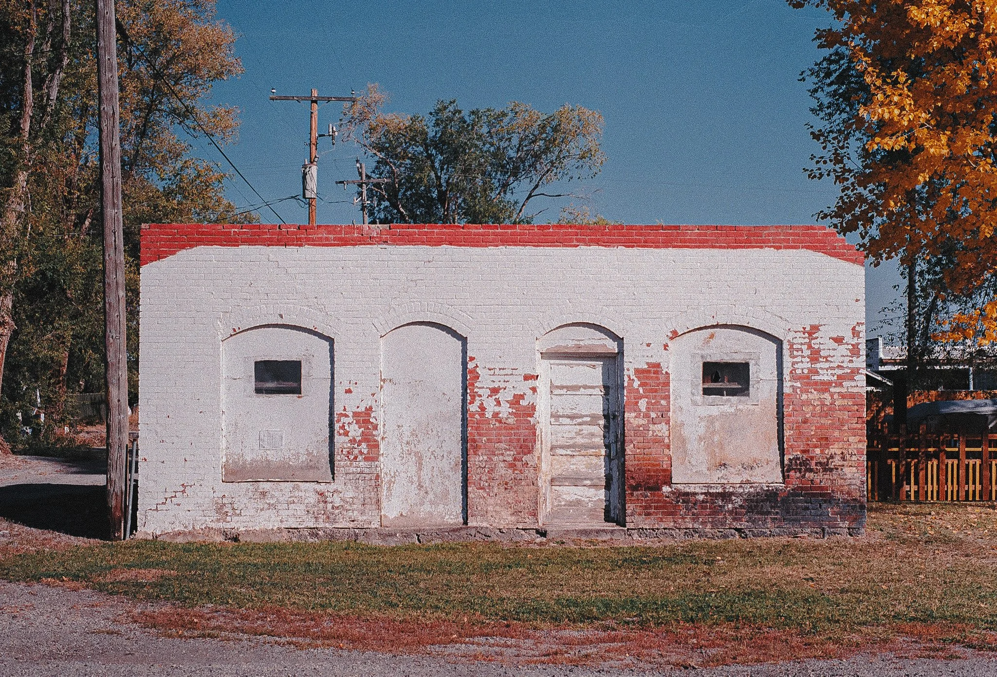 Old brick building with peeling white paint and three arched doorways, one boarded up, with small rectangular windows on each side. There are trees with autumn leaves and utility poles in the background.