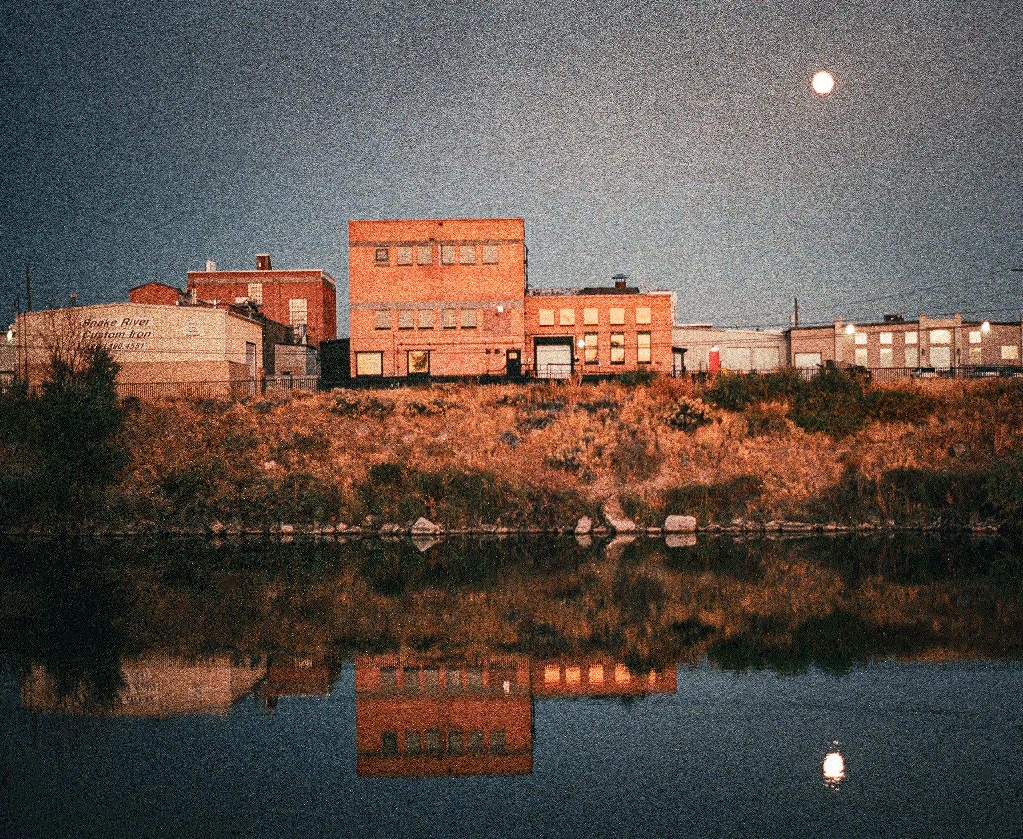Night scene of a cityscape with buildings on a hill reflected in a body of water, with a full moon in the sky.