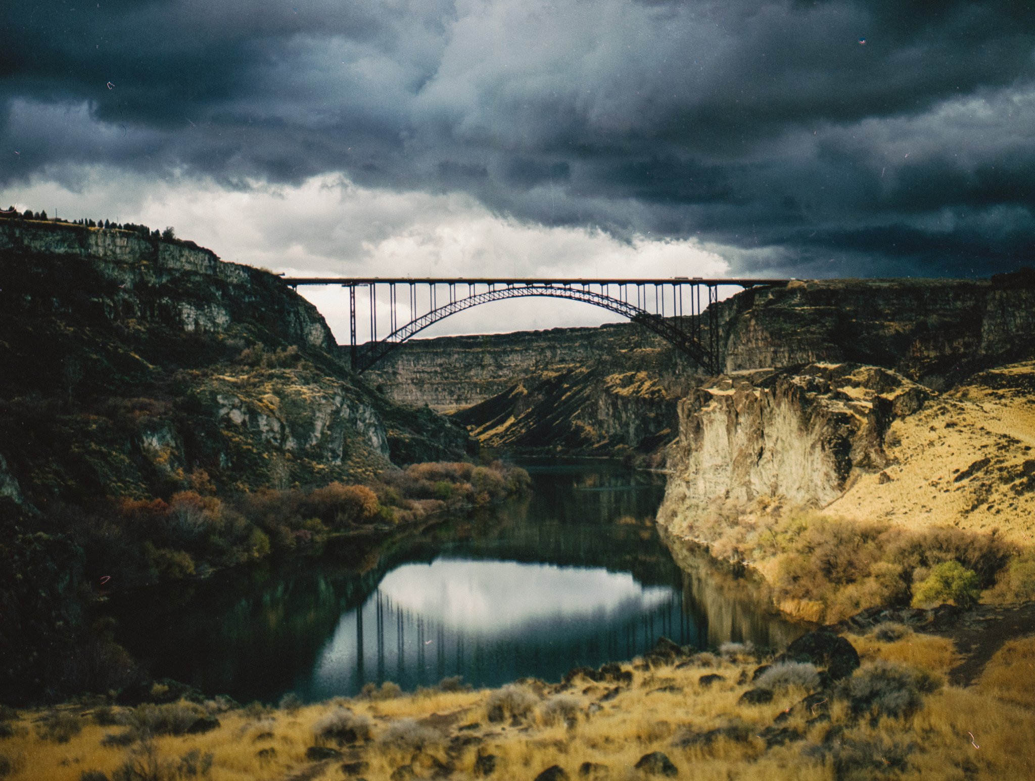 A landscape view of a canyon with a river running through it and a large arch bridge spanning the top, under a cloudy dark sky.