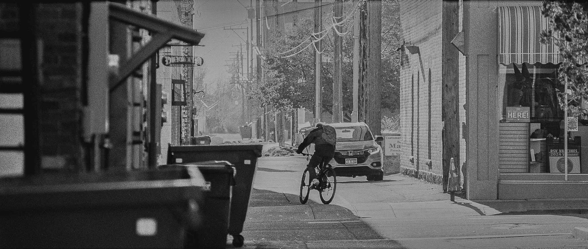 A person riding a bicycle on a quiet city street with parked cars, utility poles, and buildings, in black and white.
