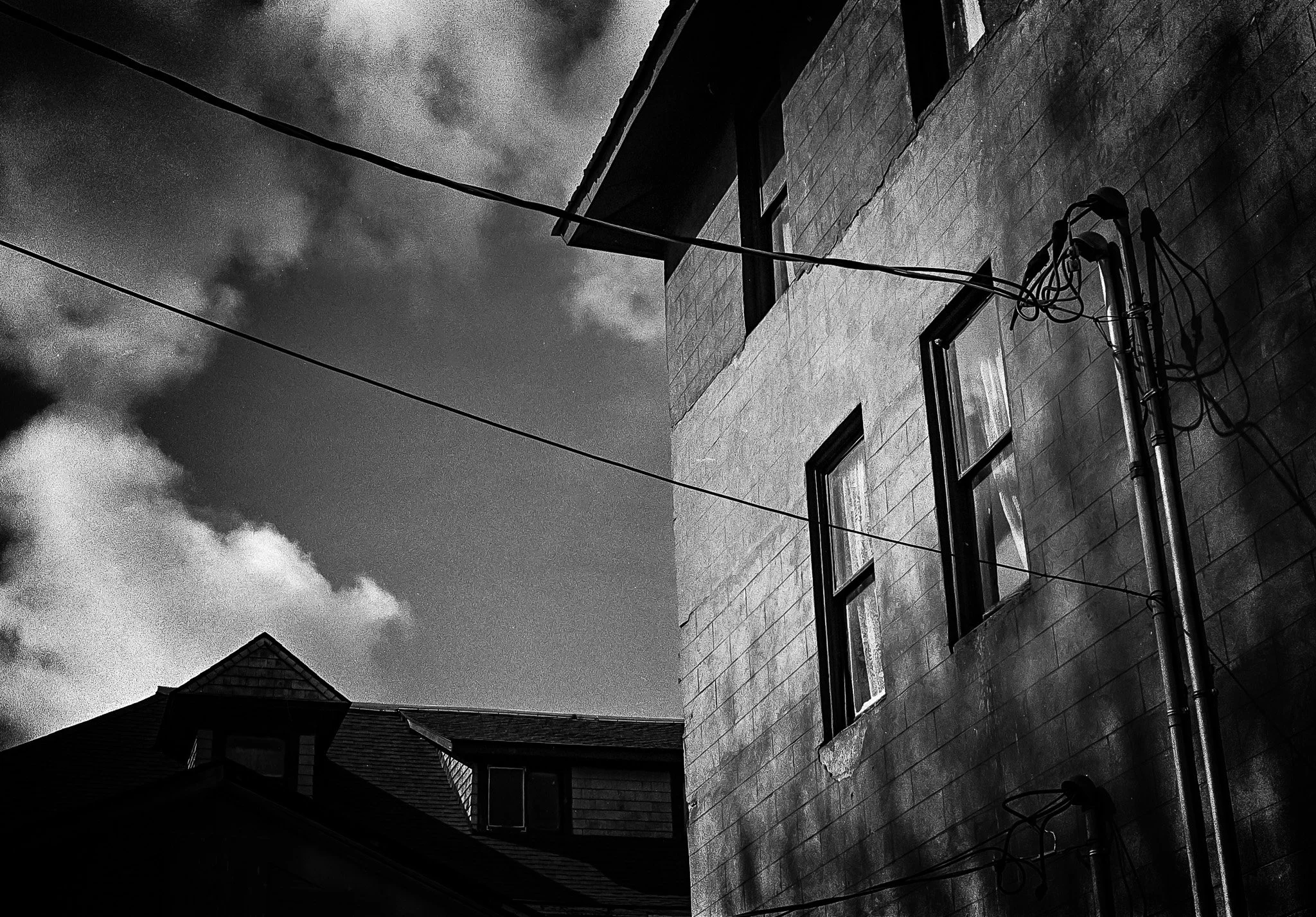 Black and white photo of an old brick building with three visible windows, a drainpipe, and some exposed wires. Part of a neighboring building's roof is also visible against a cloudy sky.