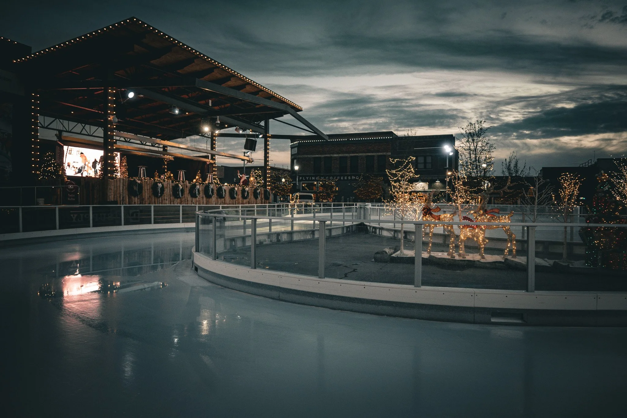 An outdoor ice skating rink decorated with Christmas lights and festive holiday figures, including reindeer, during twilight.