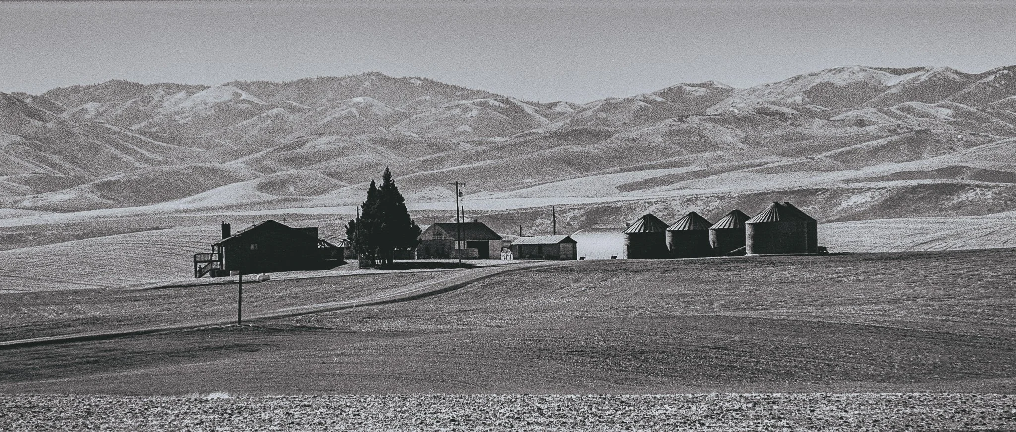 A rural farmstead with a few buildings and silos, set against rolling hills in the background, all in black and white.