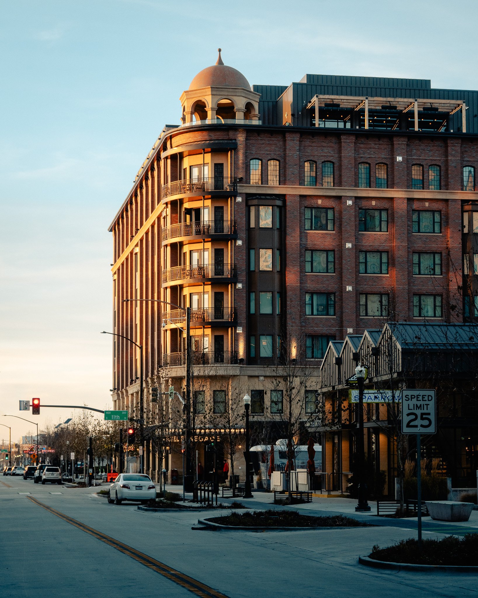 A multi-story brick building with rounded balconies and a domed tower at the corner, lining a city street at sunset with cars, street lamps, and a speed limit sign.
