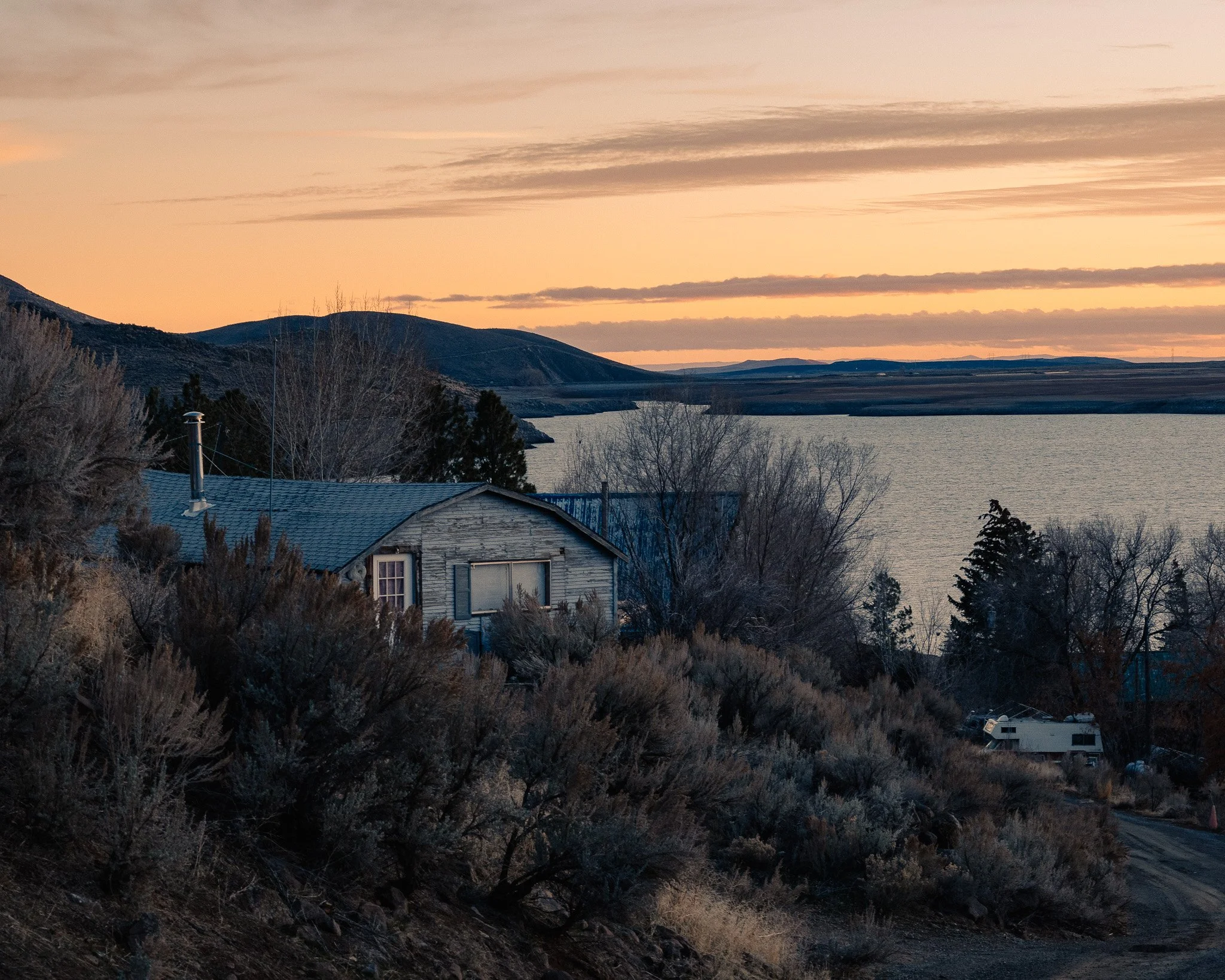 A peaceful lakeside scene at sunset, with a rustic wooden house surrounded by bushes and trees, and mountains in the distance under a partly cloudy sky.