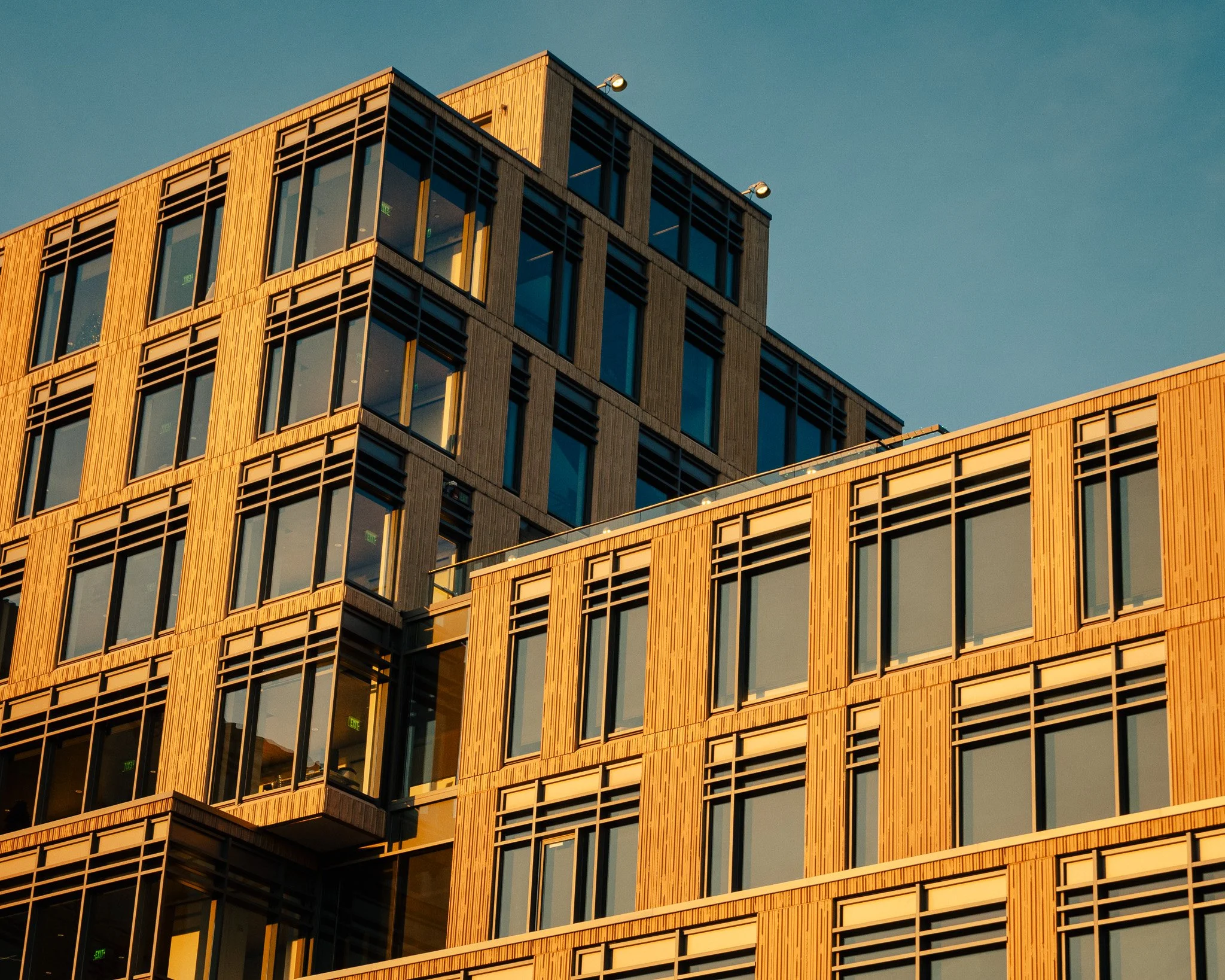 Close-up of a modern multistory building with wooden exterior panels and large glass windows, captured during sunset.
