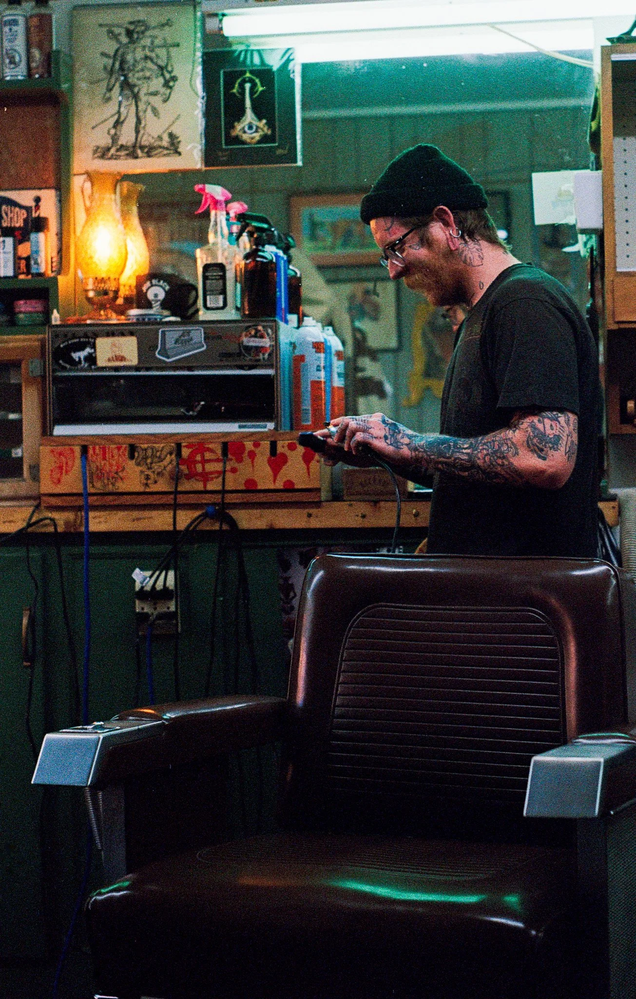 A man with tattoos, glasses, and a black beanie, smiling while looking at his phone in a barbershop or tattoo shop with various art and supplies in the background.