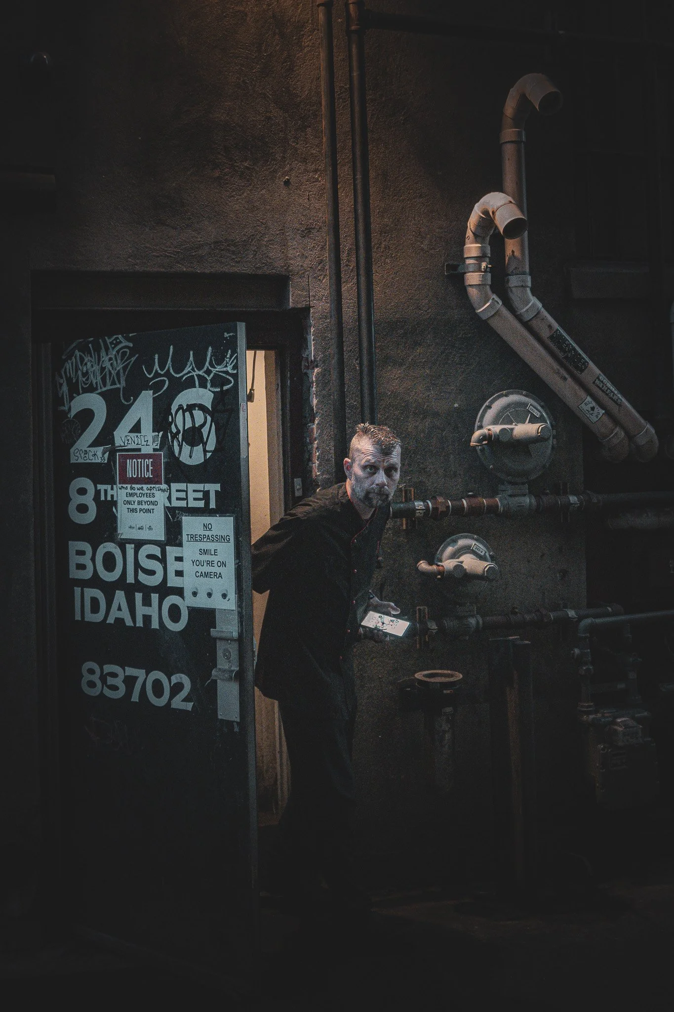A man with a beard and short hair standing in a doorway of an industrial building with numerous pipes on a dark wall, holding a small rectangular object, possibly a phone, and looking towards the camera.