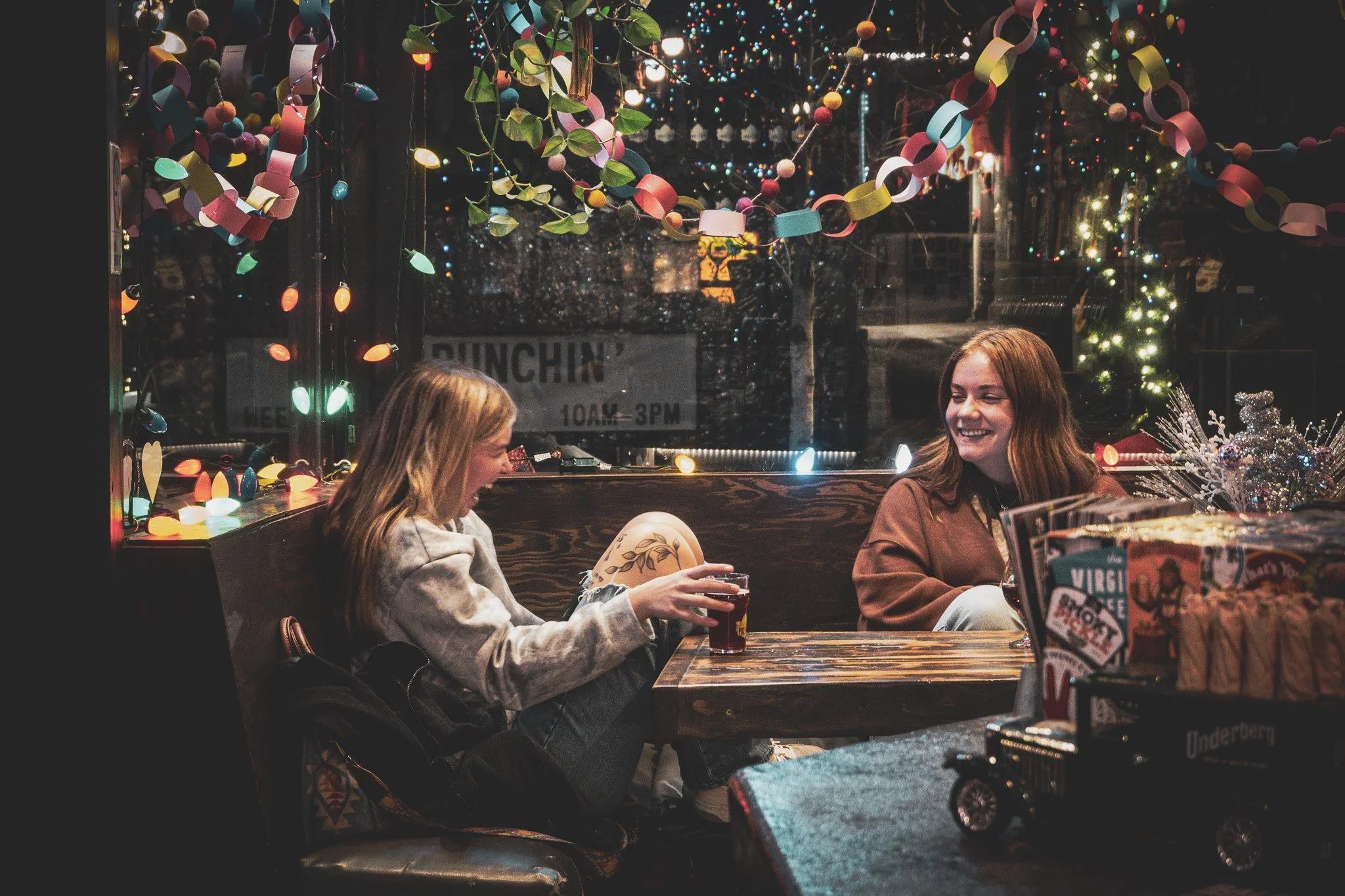 Two women sitting at a table, smiling and laughing, inside a decorated space with string lights, paper garlands, and Christmas decorations, during a festive evening.