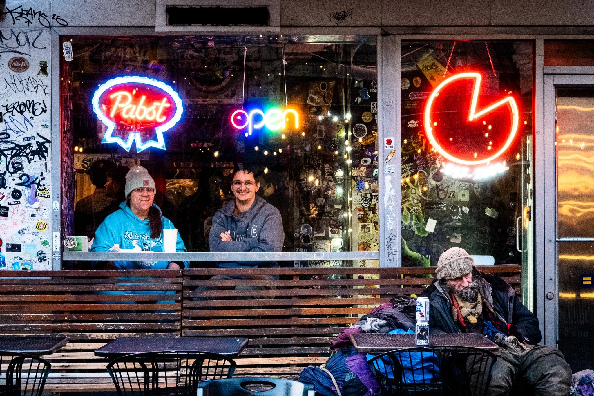 Two people are sitting outside a station chatting, with neon signs of a Pabst beer logo, an open sign, and a clock showing almost 9 o'clock hanging in the window behind them. A homeless man with a gray beanie and layered clothing is sitting on a benc